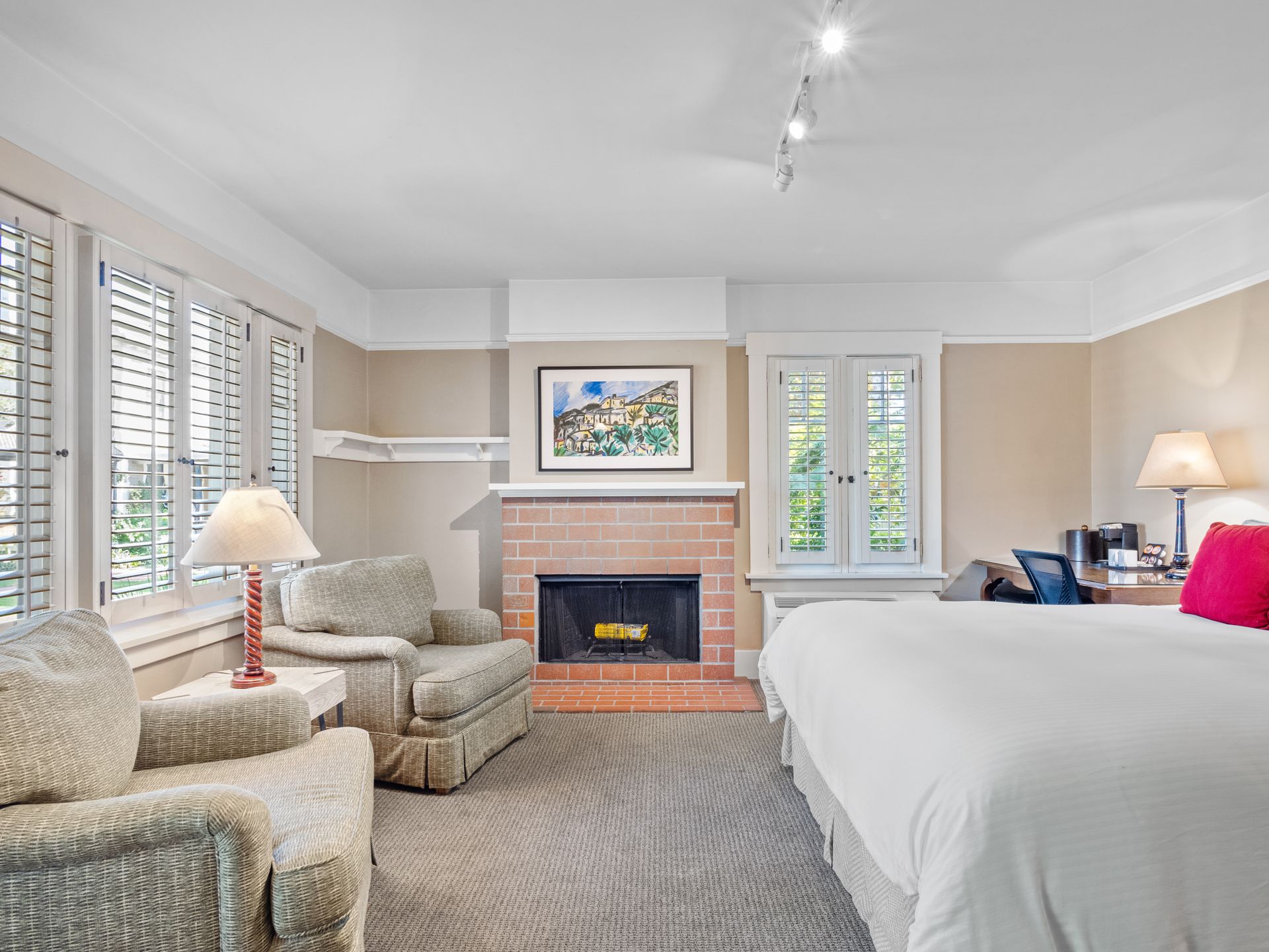 A bedroom with a king bed, two armchairs, a brick fireplace, and windows with shutters under a neutral-toned ceiling.