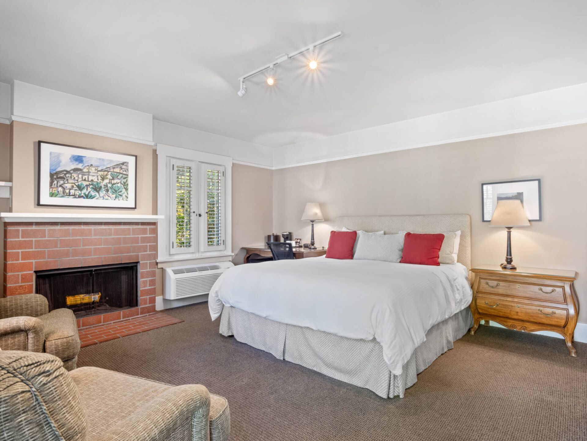A bedroom with a king-sized bed, a brick fireplace, tan walls, a bedside chest, and a desk near a window.