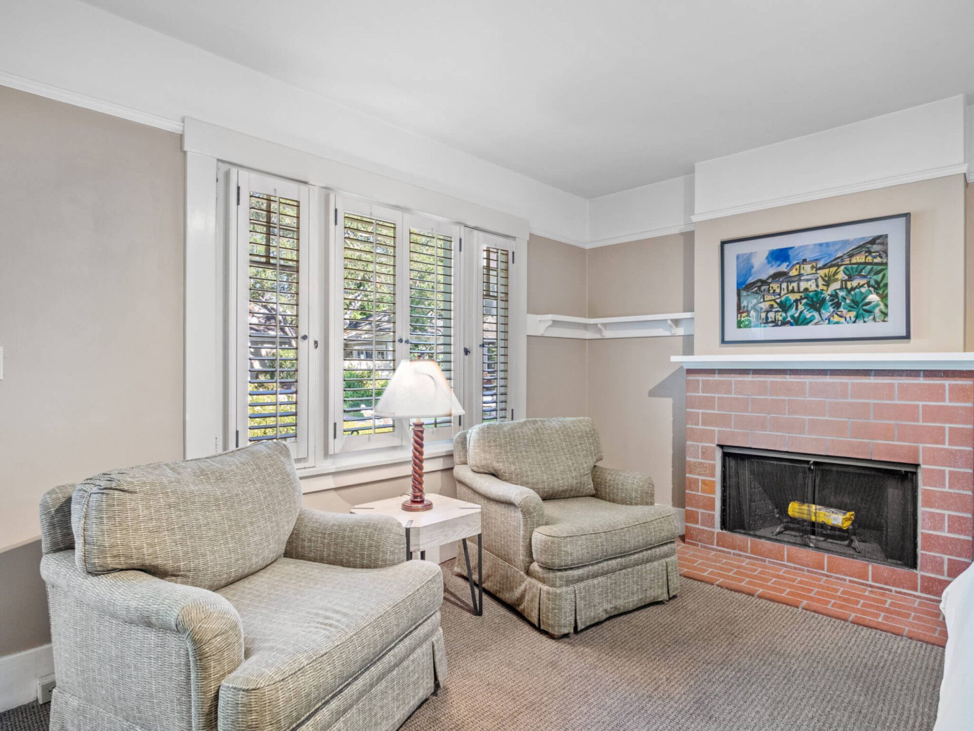 Two beige patterned armchairs sit before a brick fireplace in a sunlit room with white trim and wooden blinds.