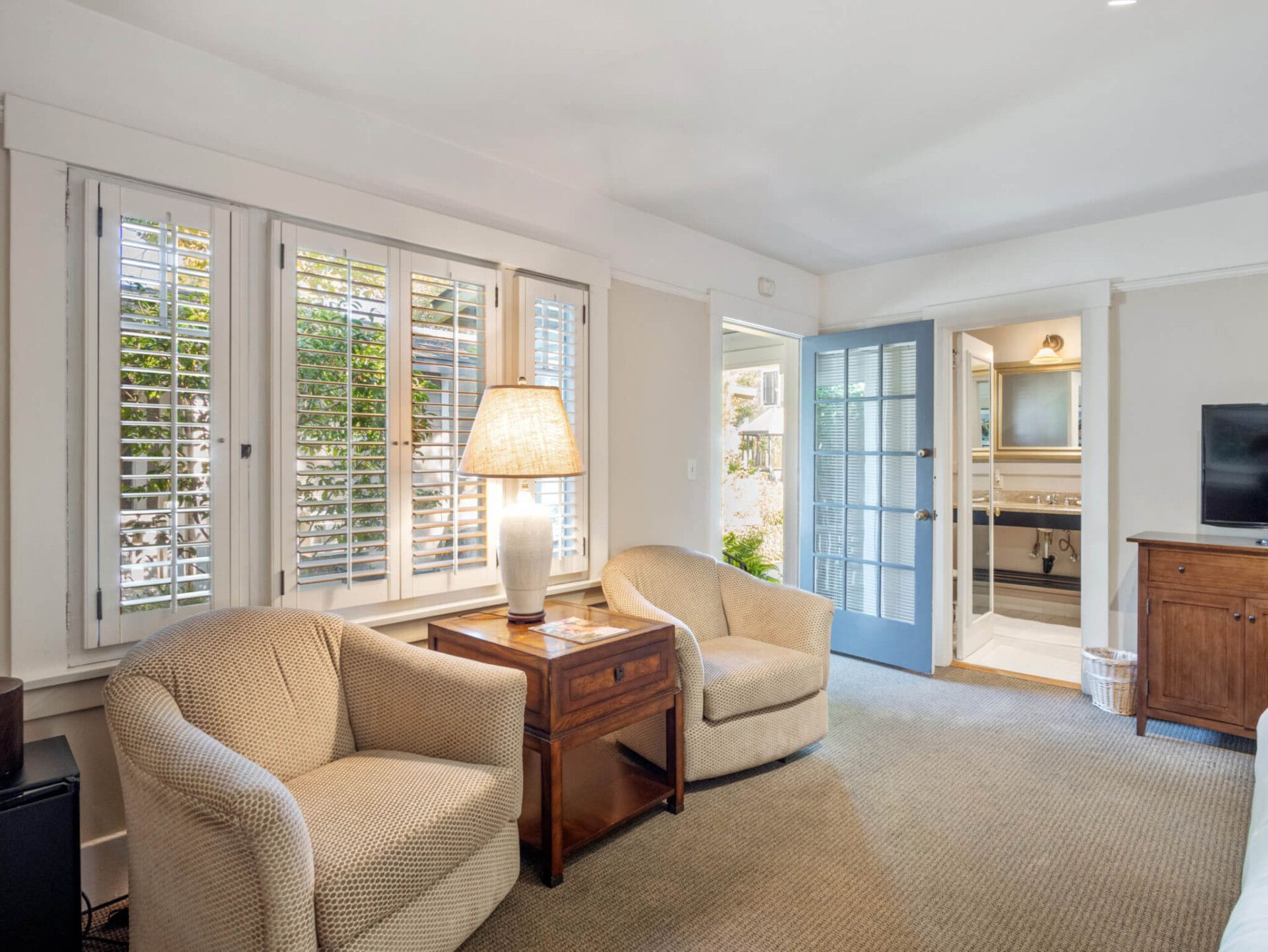 A bright living space with two beige armchairs, a wooden side table with a lamp, a blue glass door, and a view of a bathroom.