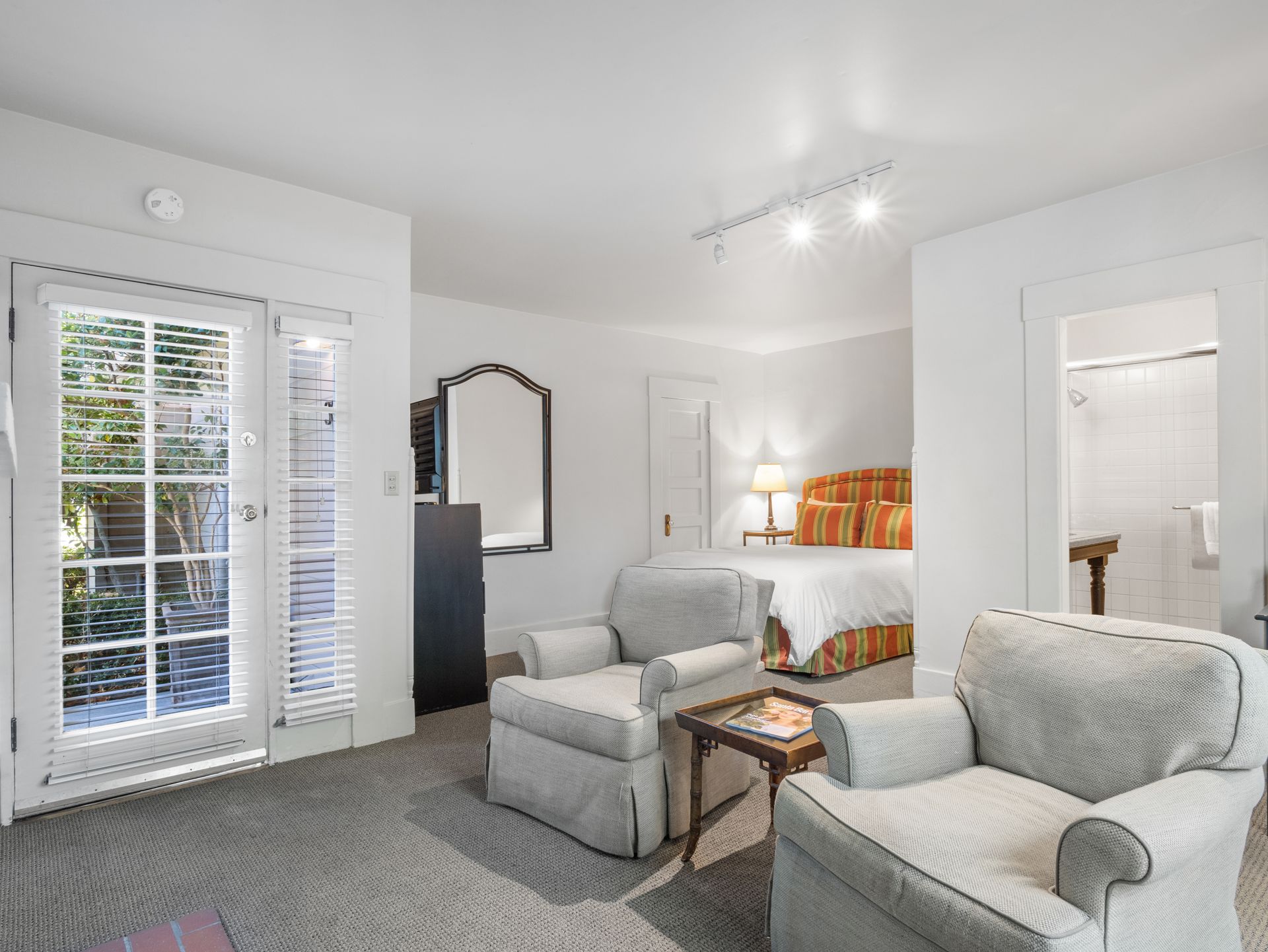 A hotel room featuring two light-colored armchairs, a bed with a gold headboard, a mirror, and a door to a small patio.