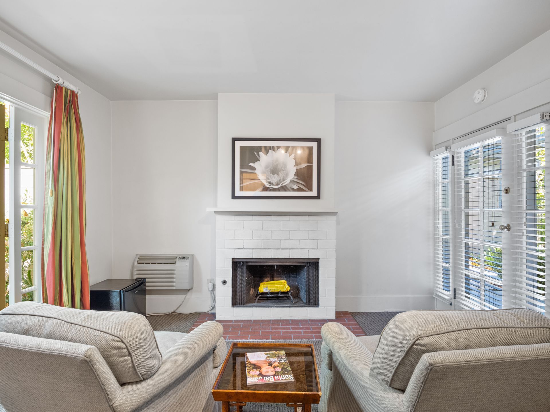 A bright living room with two neutral-colored chairs, a small coffee table, and a centered white brick fireplace.