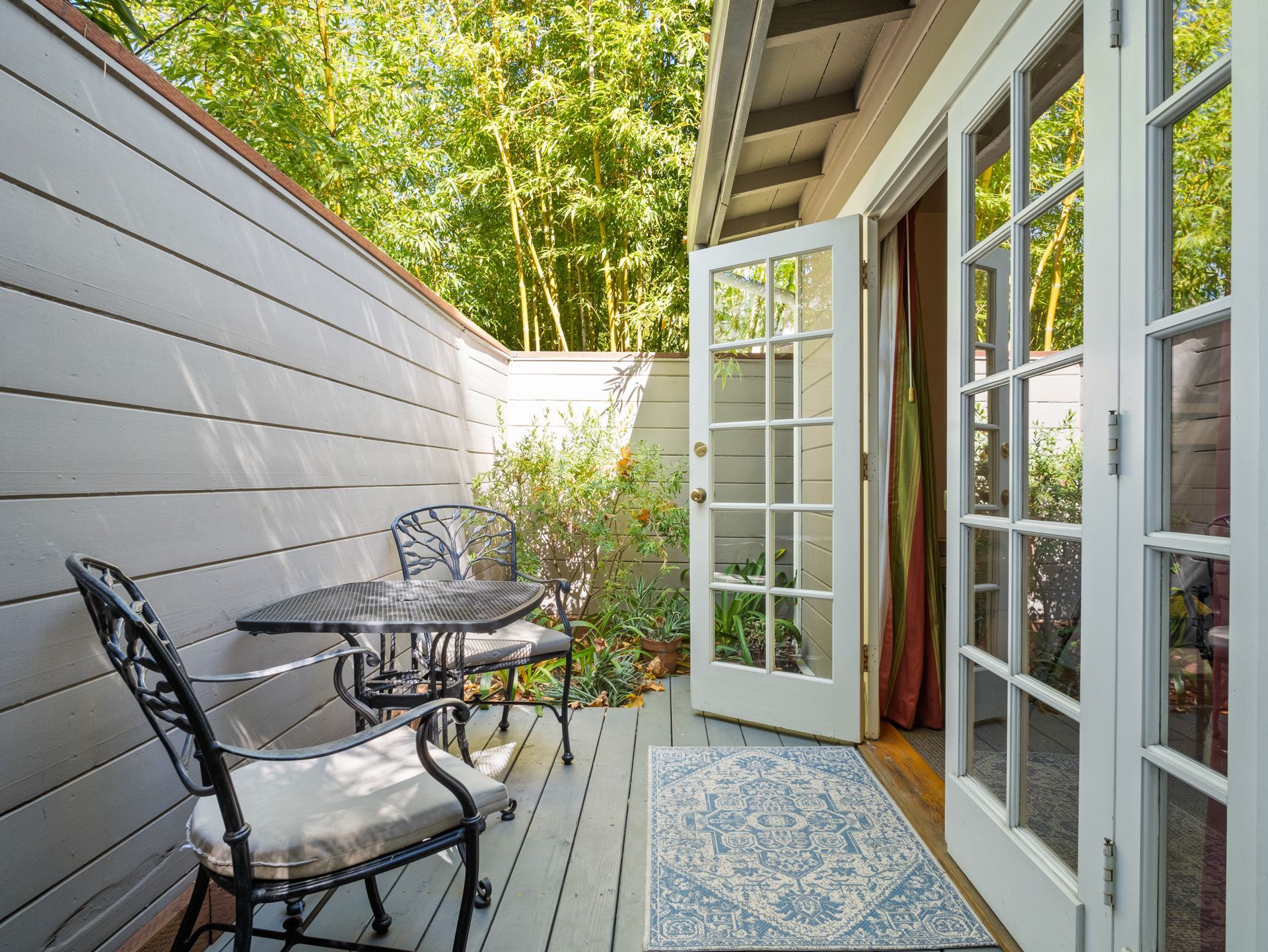 A patio with a metal table and two chairs, next to white French doors leading inside, with a small patterned rug.