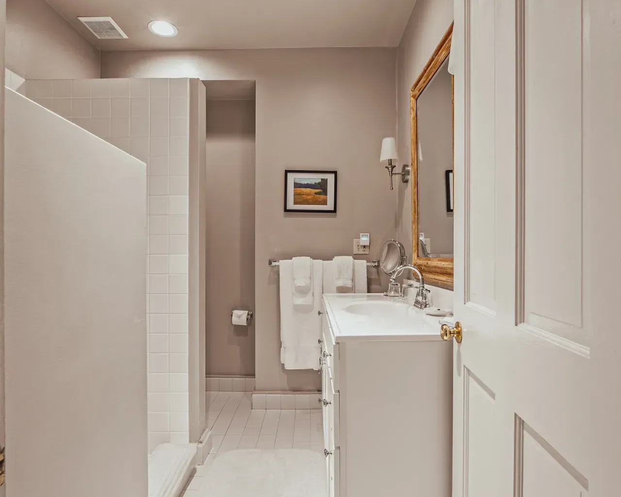 A beige bathroom featuring a white vanity with a gold-framed mirror, a walk-in tiled shower, and towels on a rack.