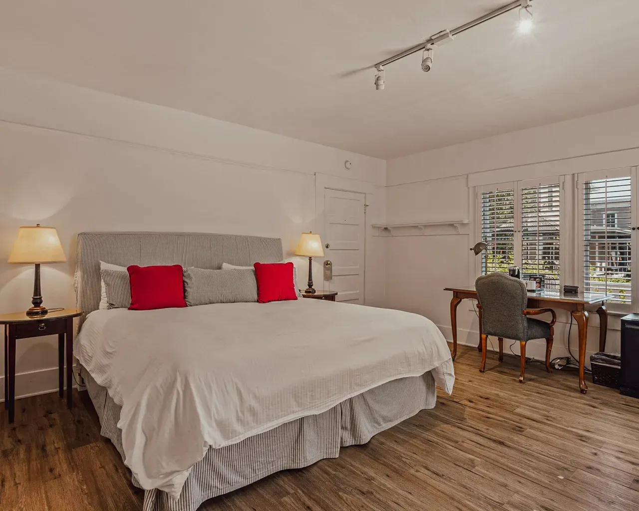 A bedroom with a king-sized bed featuring a striped headboard and red pillows, a desk, and windows with white shutters.