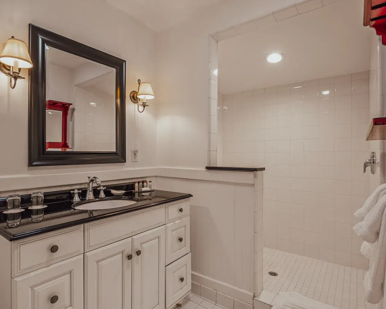 A bathroom with a white vanity, black granite countertop, dark framed mirror, and a walk-in shower with white tiled walls.
