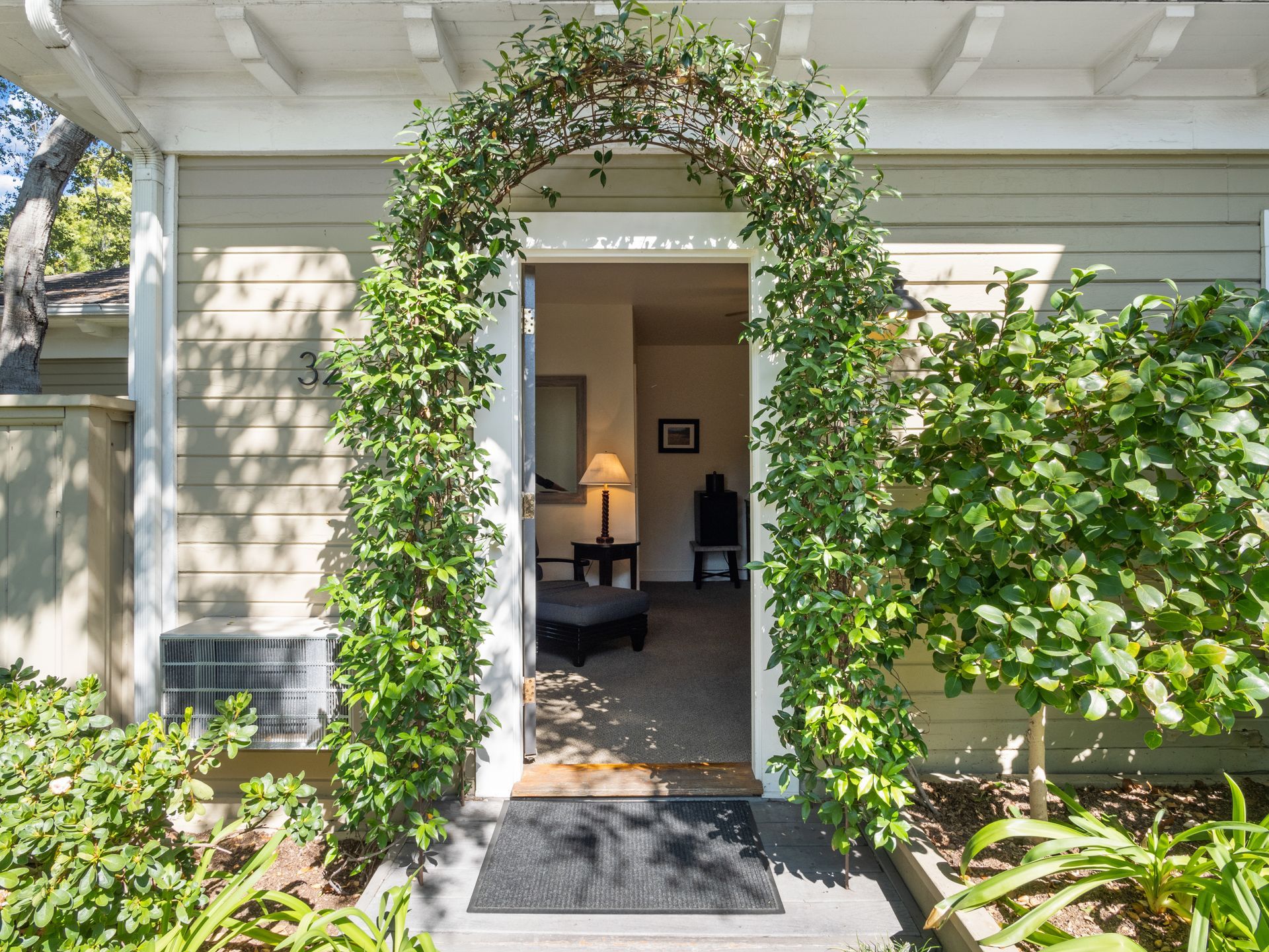 A front entryway framed by a leafy green archway, leading into a sunlit room with a table lamp and furniture.