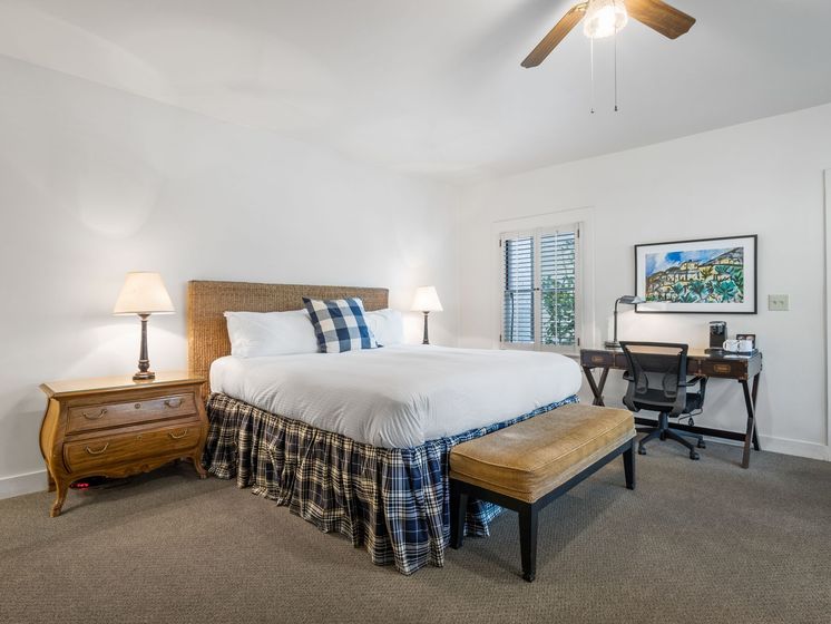 A hotel-style bedroom features a bed with a woven headboard, plaid bed skirt, nightstand, desk, and landscape art.