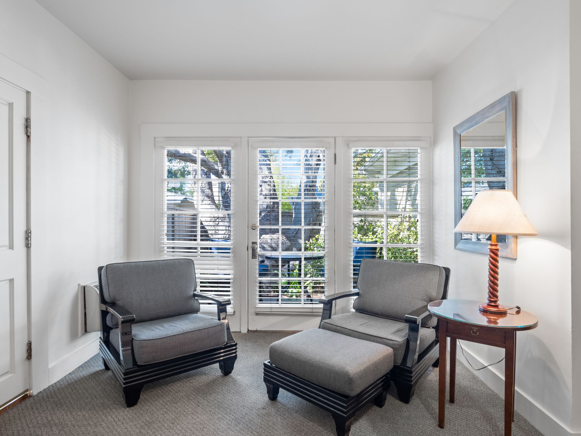 Two grey armchairs, one with an ottoman, sit near a windowed door and a side table with a lamp in a white-walled room.