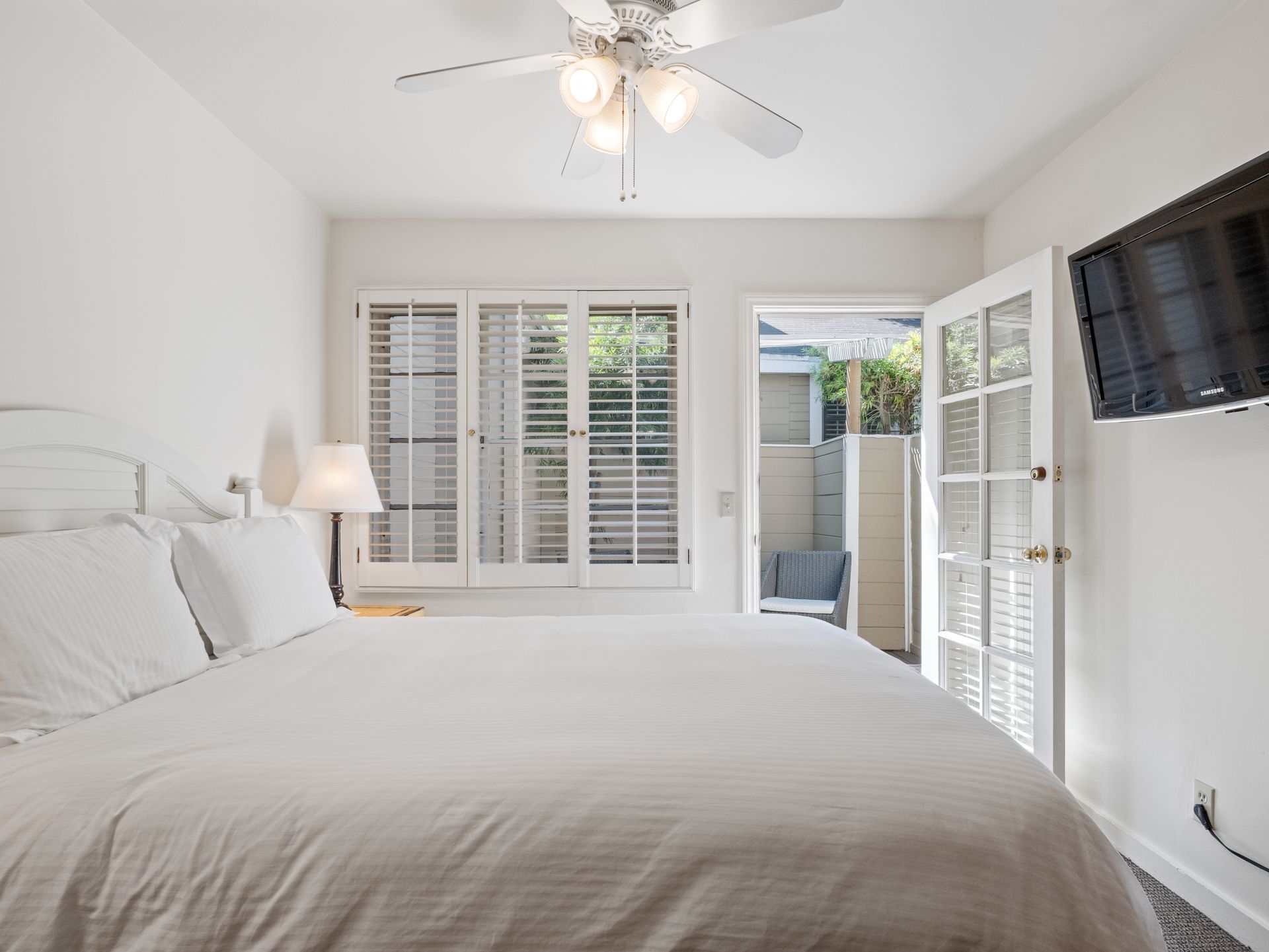 A white bedroom with a bed, ceiling fan, plantation shutters, and a television mounted on the wall near a patio door.