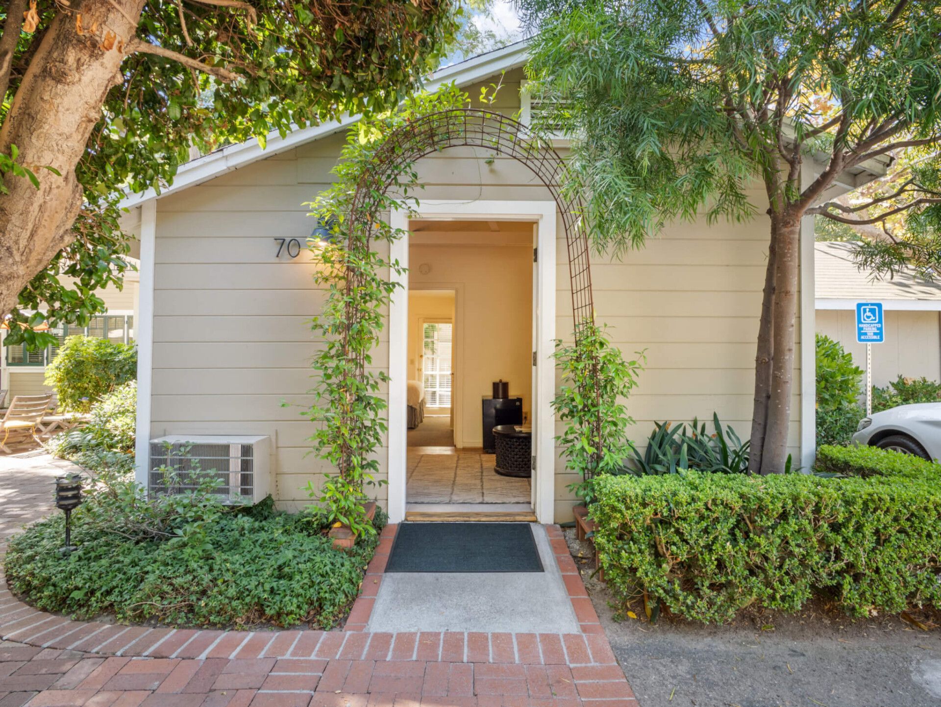 A tan building with a white-trimmed doorway, arched trellis with vines, brick walkway, and green landscaping.