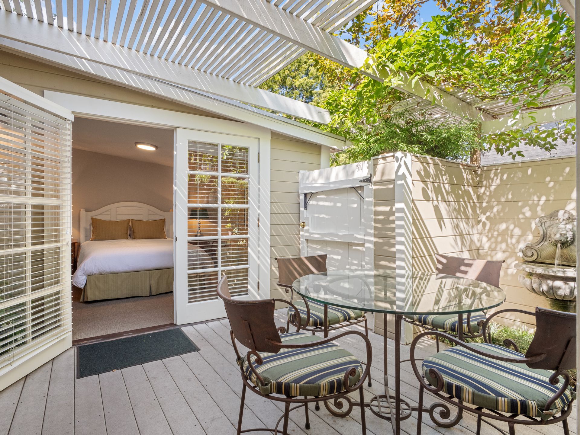 A patio with a glass-topped table and chairs leads into a bedroom through open French doors under a pergola.