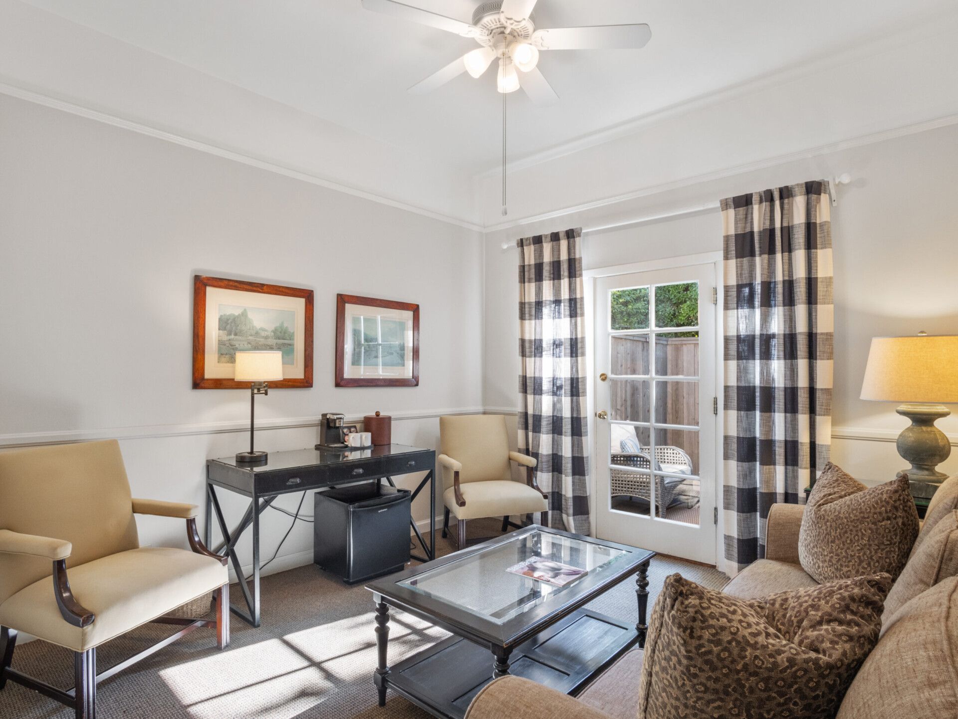 A bright, neutral-toned living room featuring tan armchairs, a glass coffee table, a dark desk, and plaid curtains.