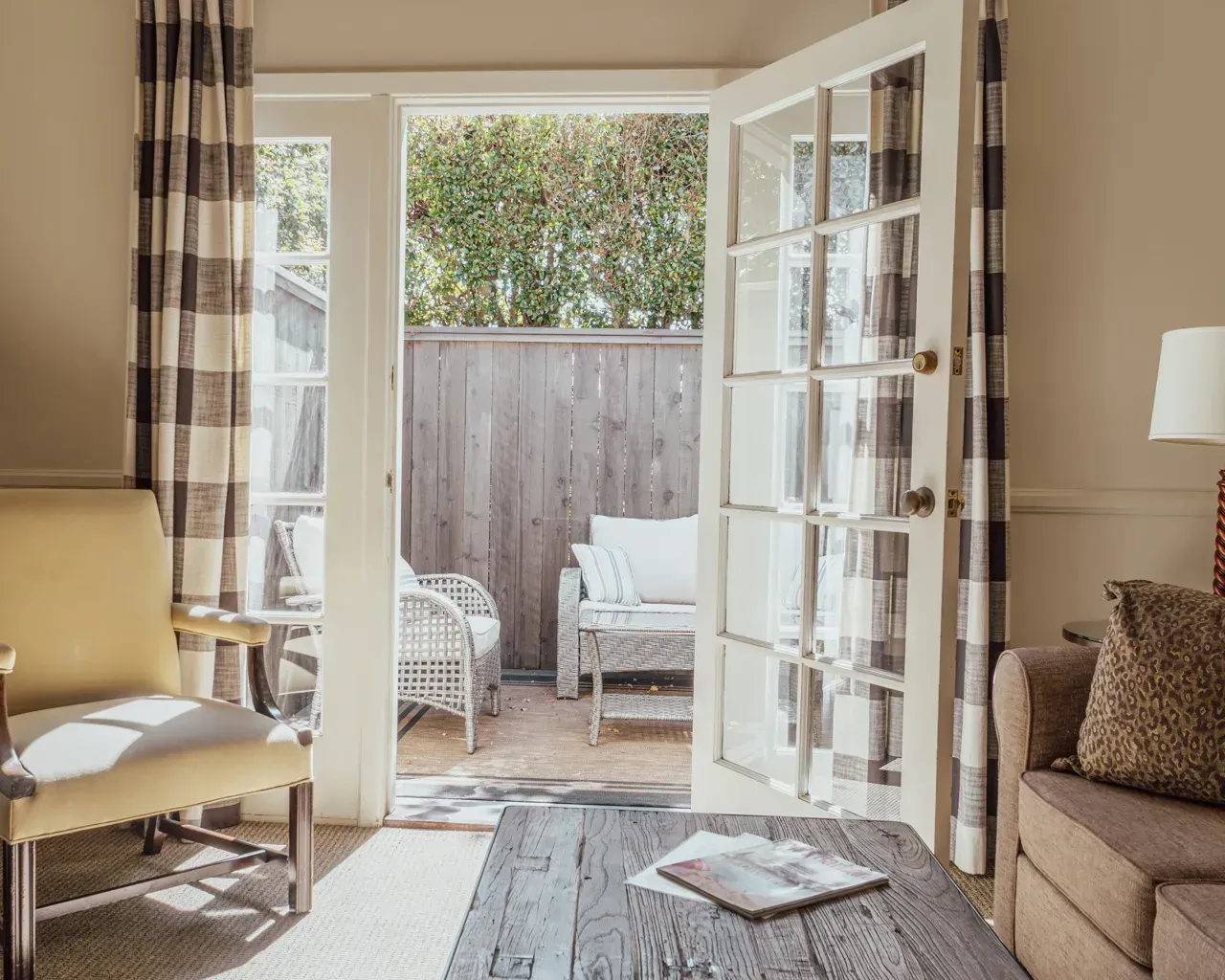 A cozy living room opens to a sunny patio with white wicker chairs and a wood fence through a glass French door.