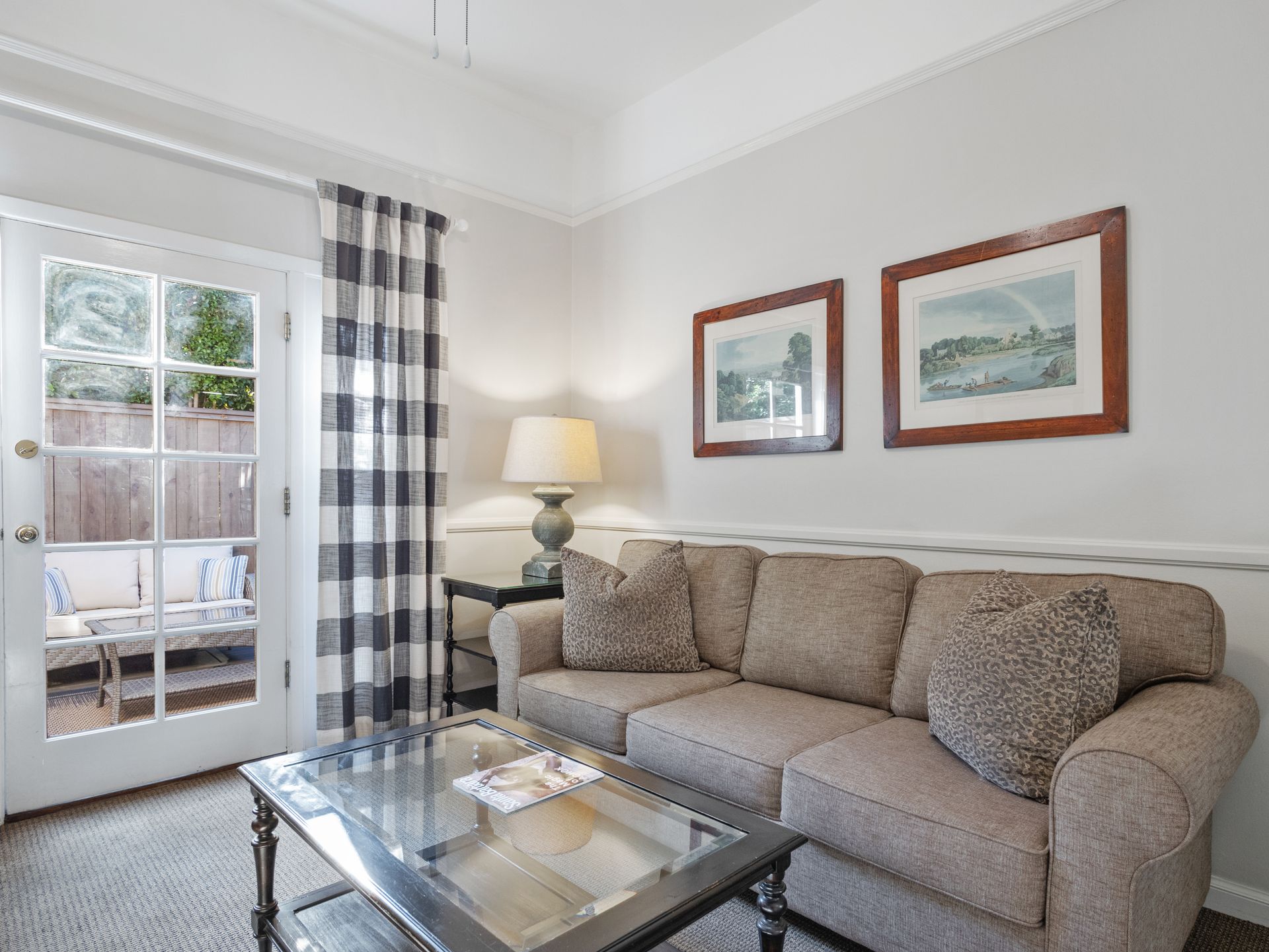 A cozy living room with a beige sofa, coffee table, lamp, and glass door leading to a patio with outdoor furniture.