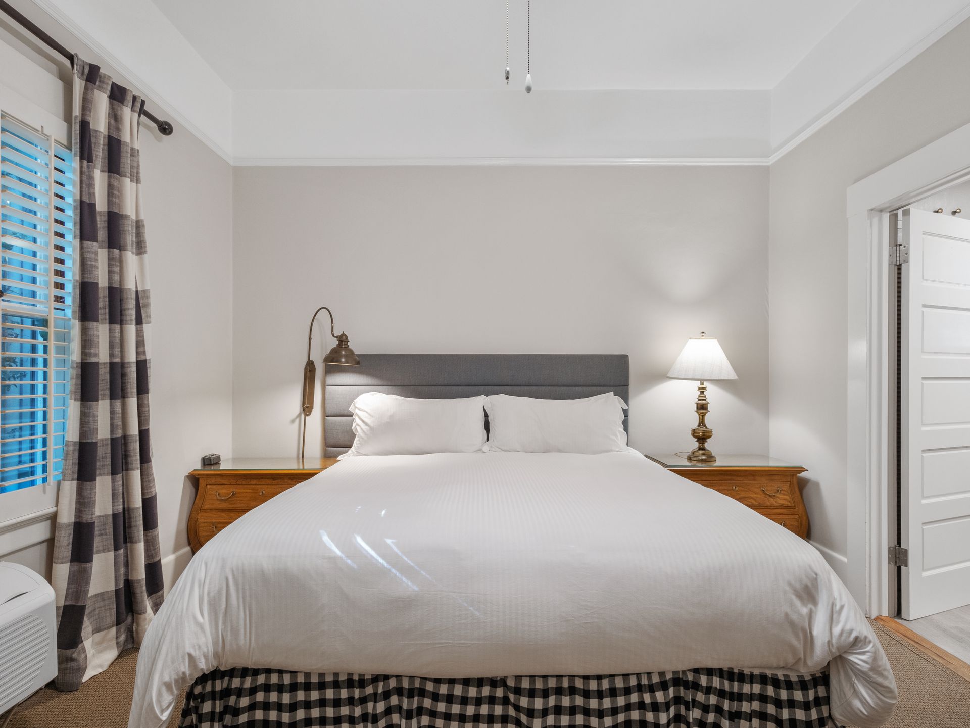 A bedroom featuring a queen bed with white bedding, a gray headboard, two wooden nightstands, and plaid-patterned accents.