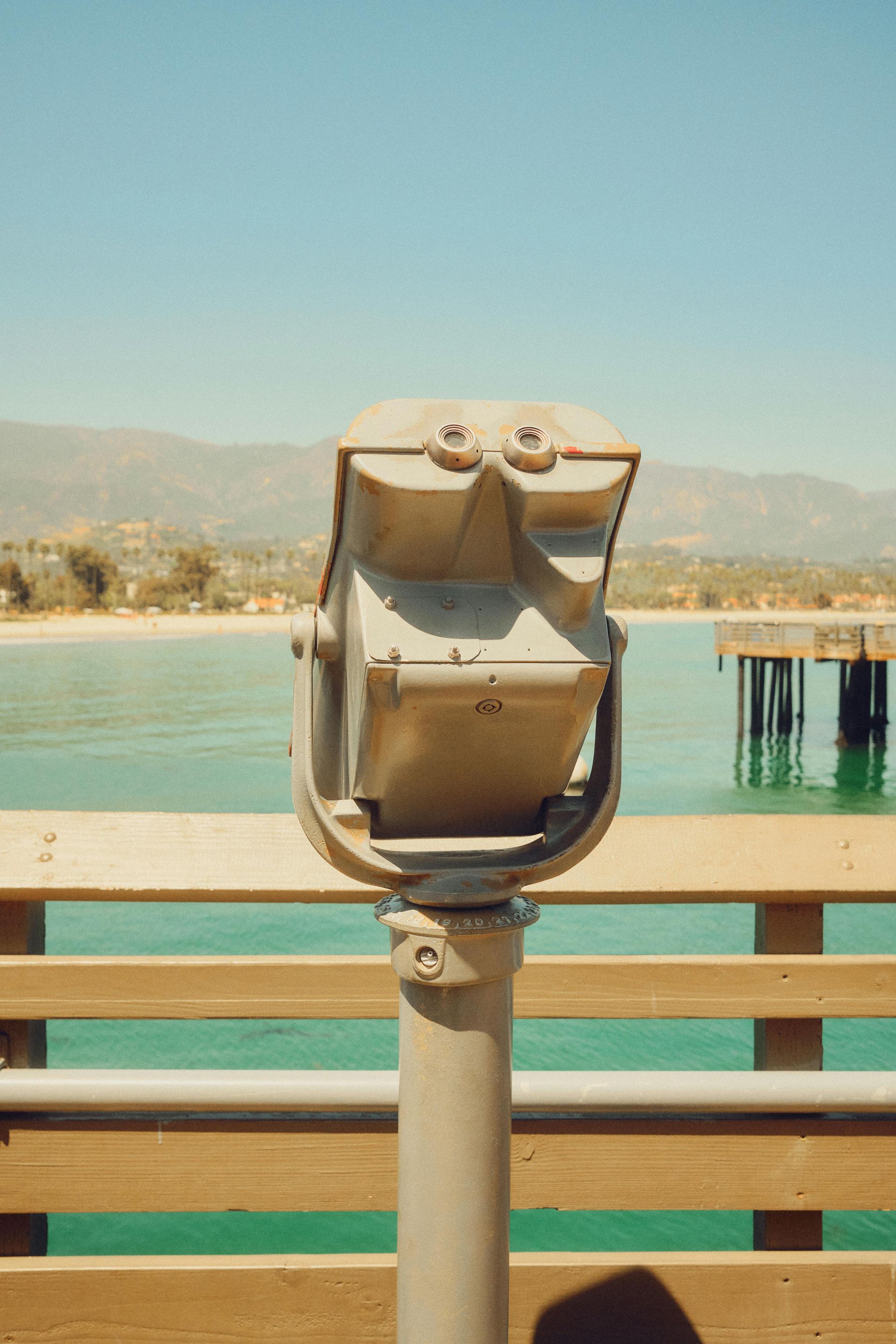 A stationary coin-operated telescope overlooks a turquoise ocean and pier on a sunny day.