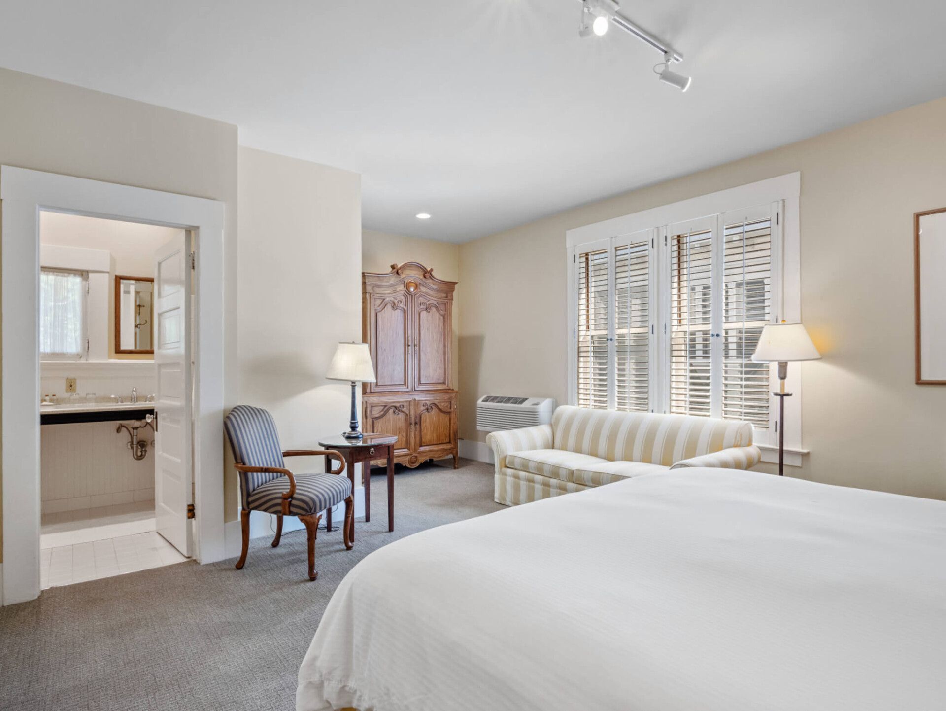 A bright bedroom with a large bed, a striped sofa, an armchair, an armoire, and an open doorway to a bathroom.