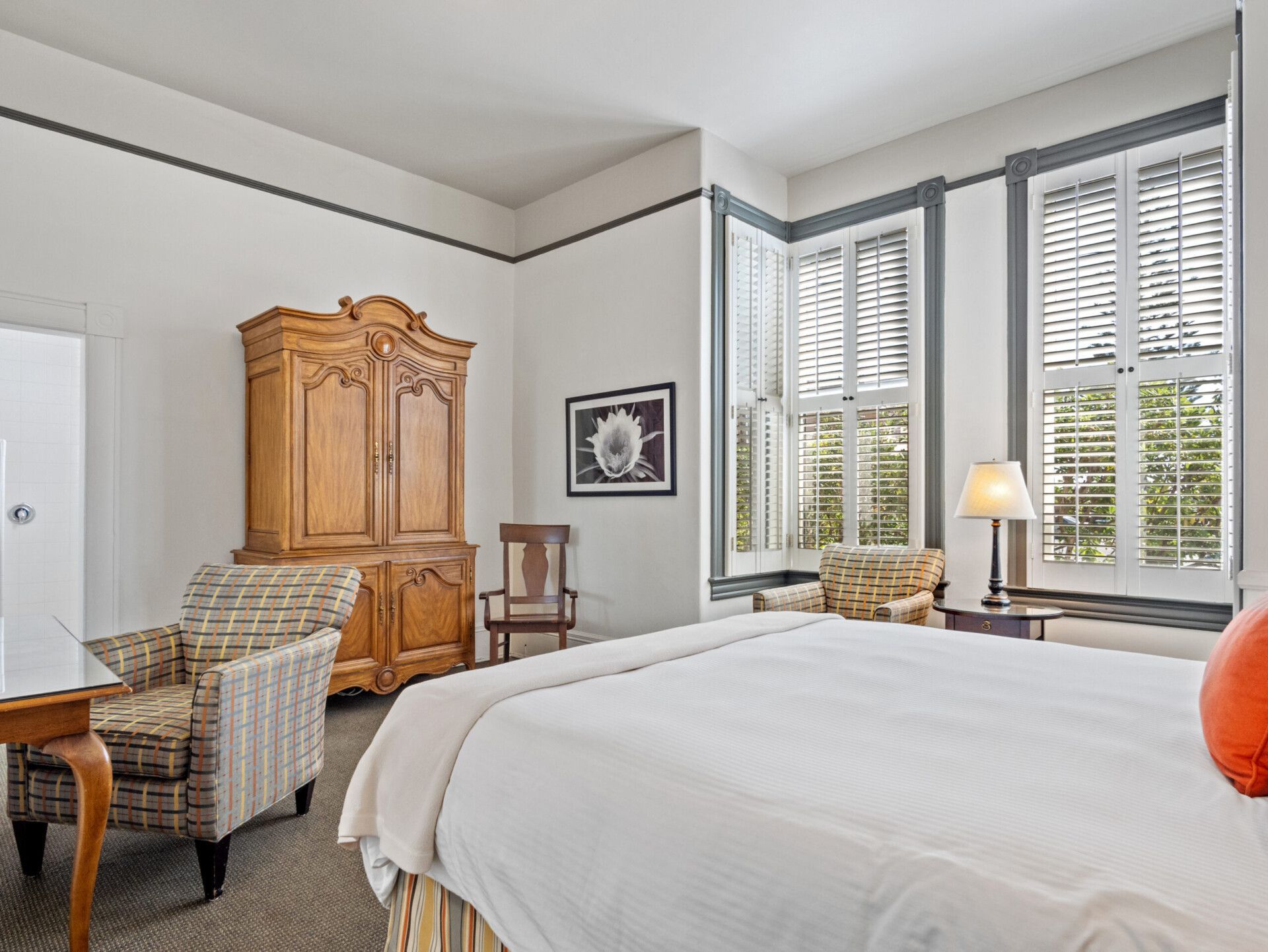 A bedroom featuring a large bed, patterned armchair, wooden armoire, and two windows with white plantation shutters.