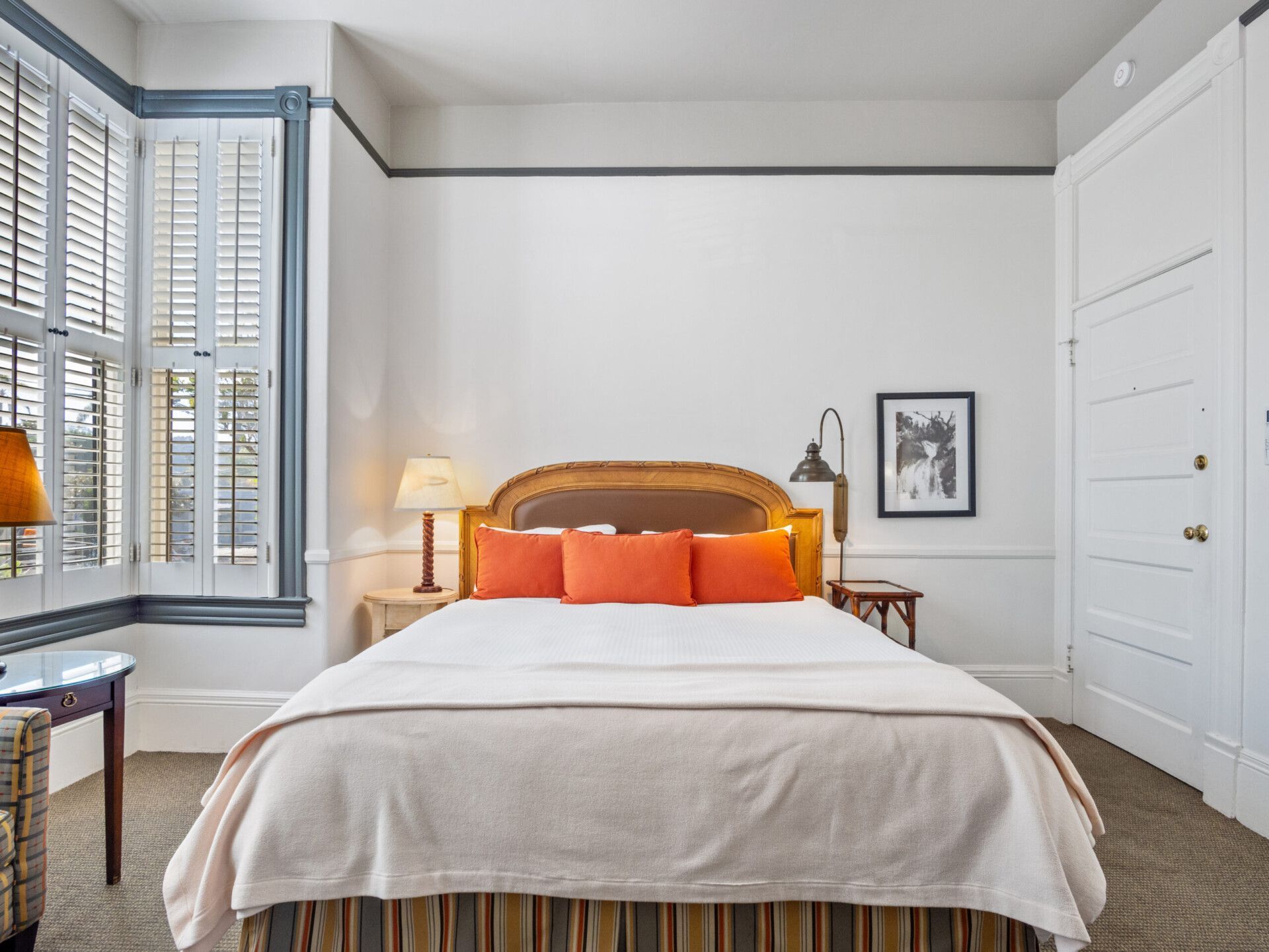 A bedroom featuring a queen bed with orange pillows, light bedding, a bedside lamp, and a window with plantation shutters.