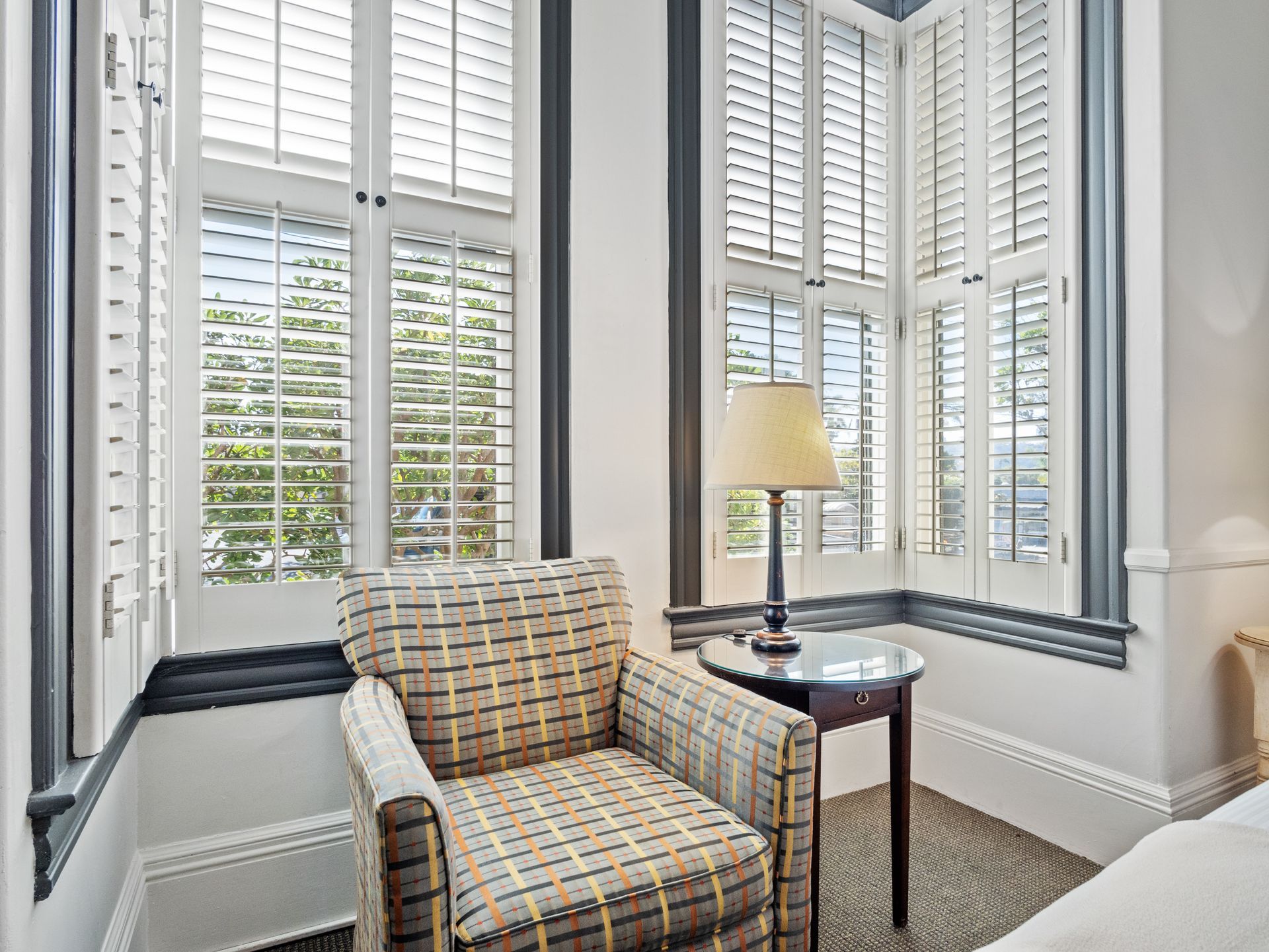 A plaid armchair sits next to a small side table with a lamp, positioned in a corner with white plantation shutters.