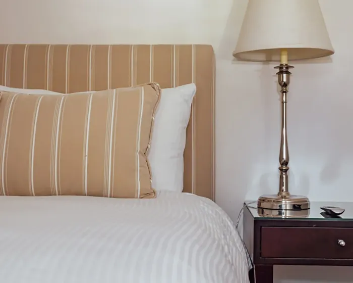 A bedroom scene featuring a striped beige headboard, a matching pillow, a white bedspread, and a lamp on a bedside table.