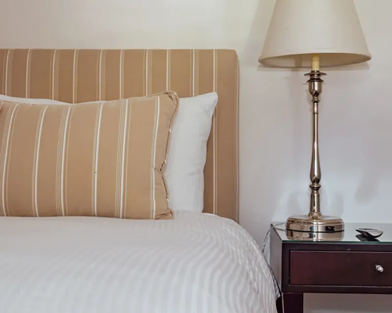 A bedroom scene featuring a striped beige headboard, a matching pillow, a white bedspread, and a lamp on a bedside table.