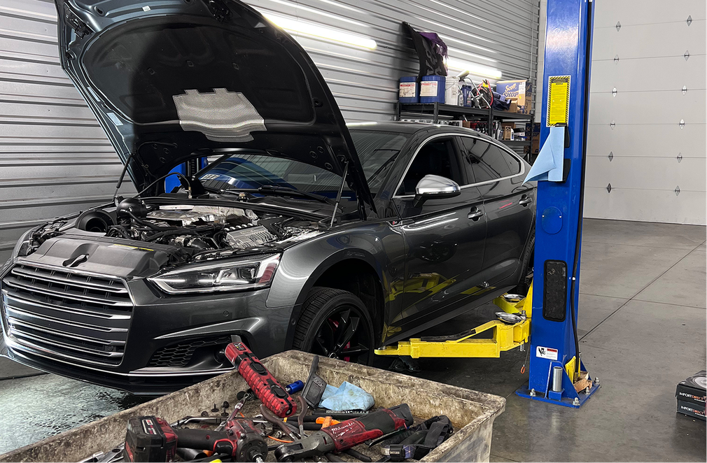 Gray car with open hood on a lift inside a garage; tools in a bin in front. | Mike's Garage CC