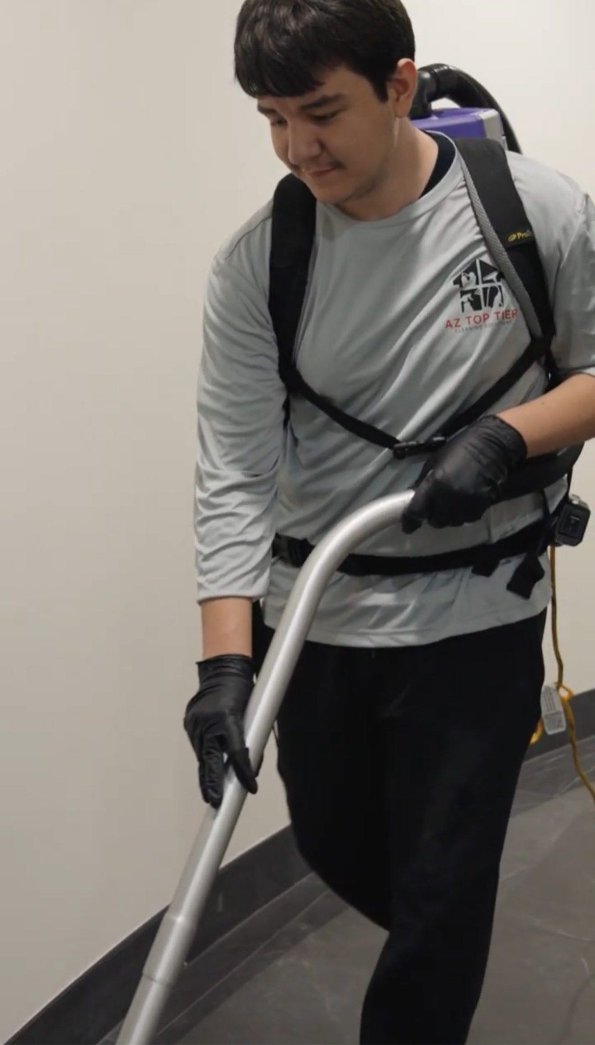 Person wearing a backpack vacuum, cleaning a hallway. They are wearing black gloves and a gray shirt.