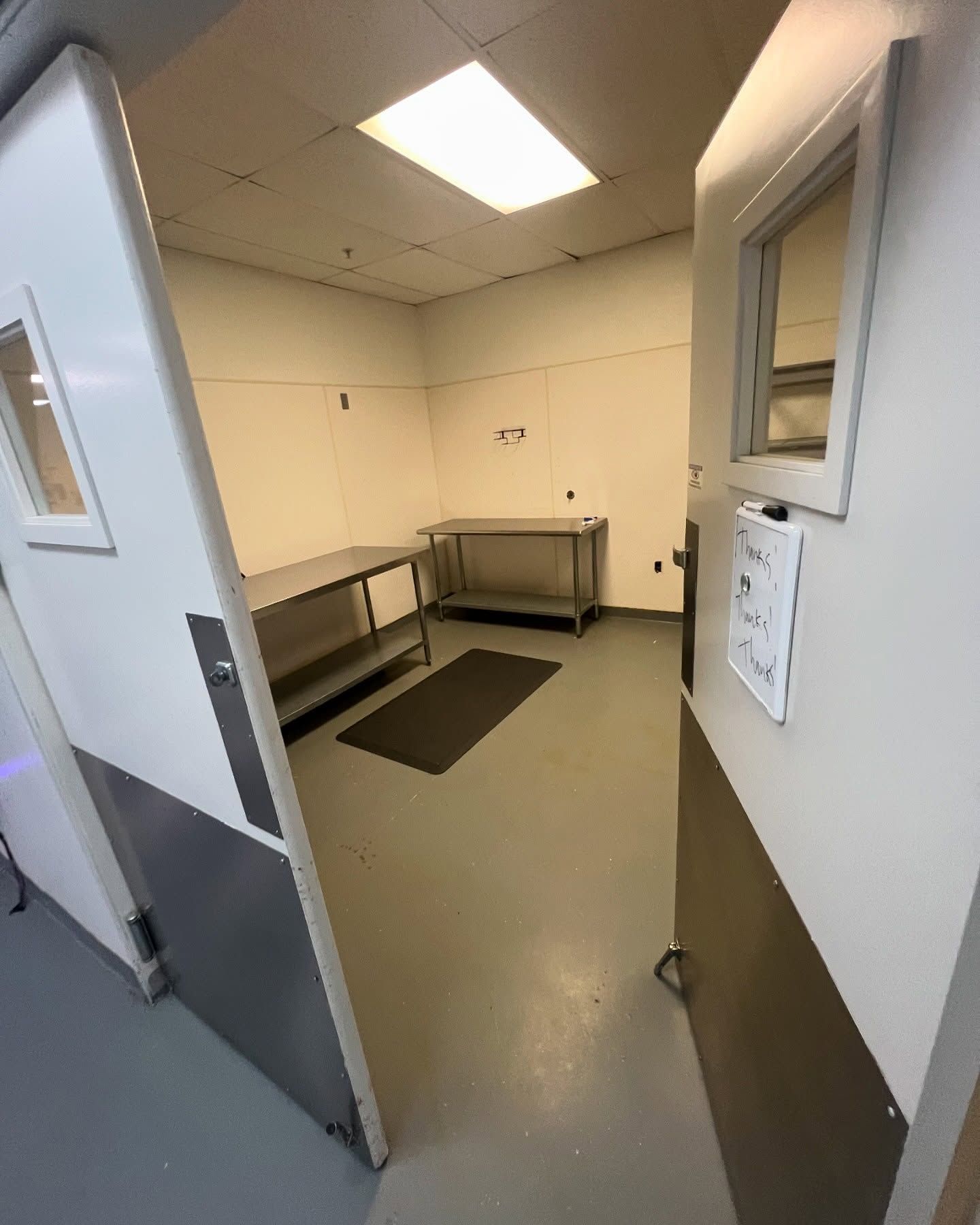 Open doorway into a sterile room with metal tables and a dark floor mat; a fluorescent light is overhead.