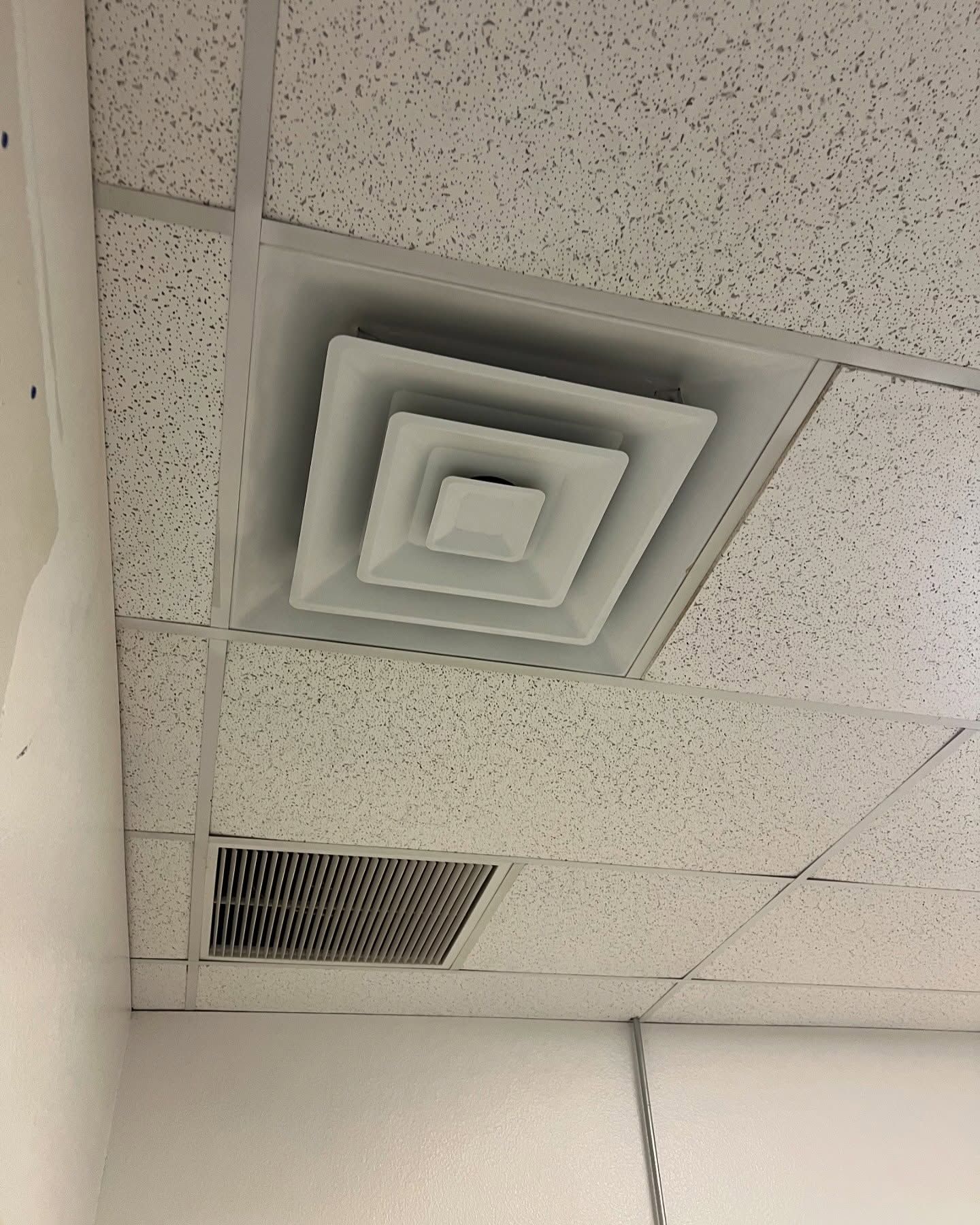 White air vent in a drop ceiling; below is a dark vent grate near a white wall.