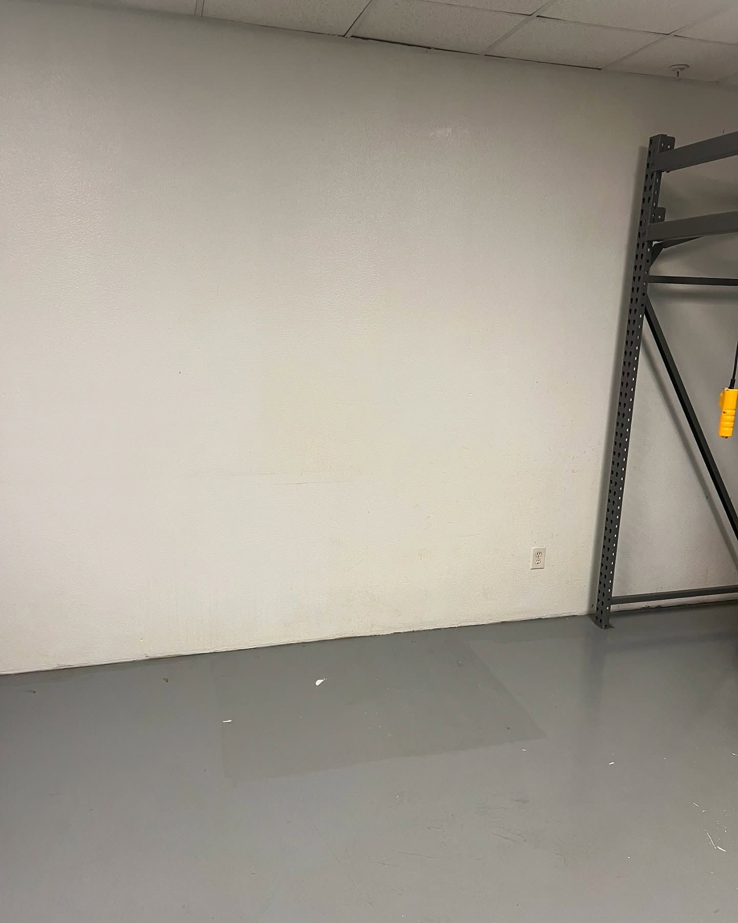 White wall in a room with a gray floor and metal shelving on the right side.