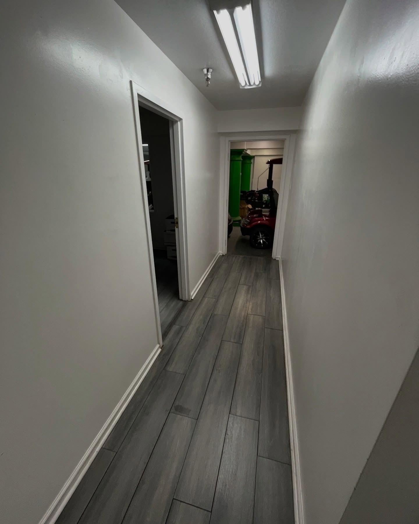 Narrow hallway with gray wood-look flooring and white walls, leading to a doorway with equipment.