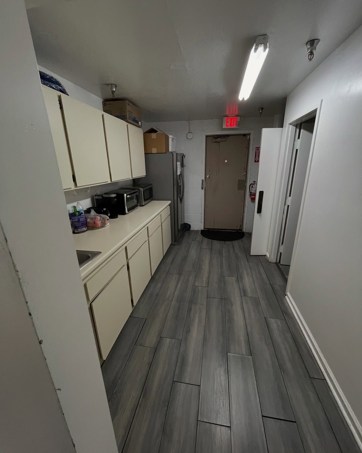 Narrow hallway with cabinets, appliances, and an exit door. Gray flooring and white walls.