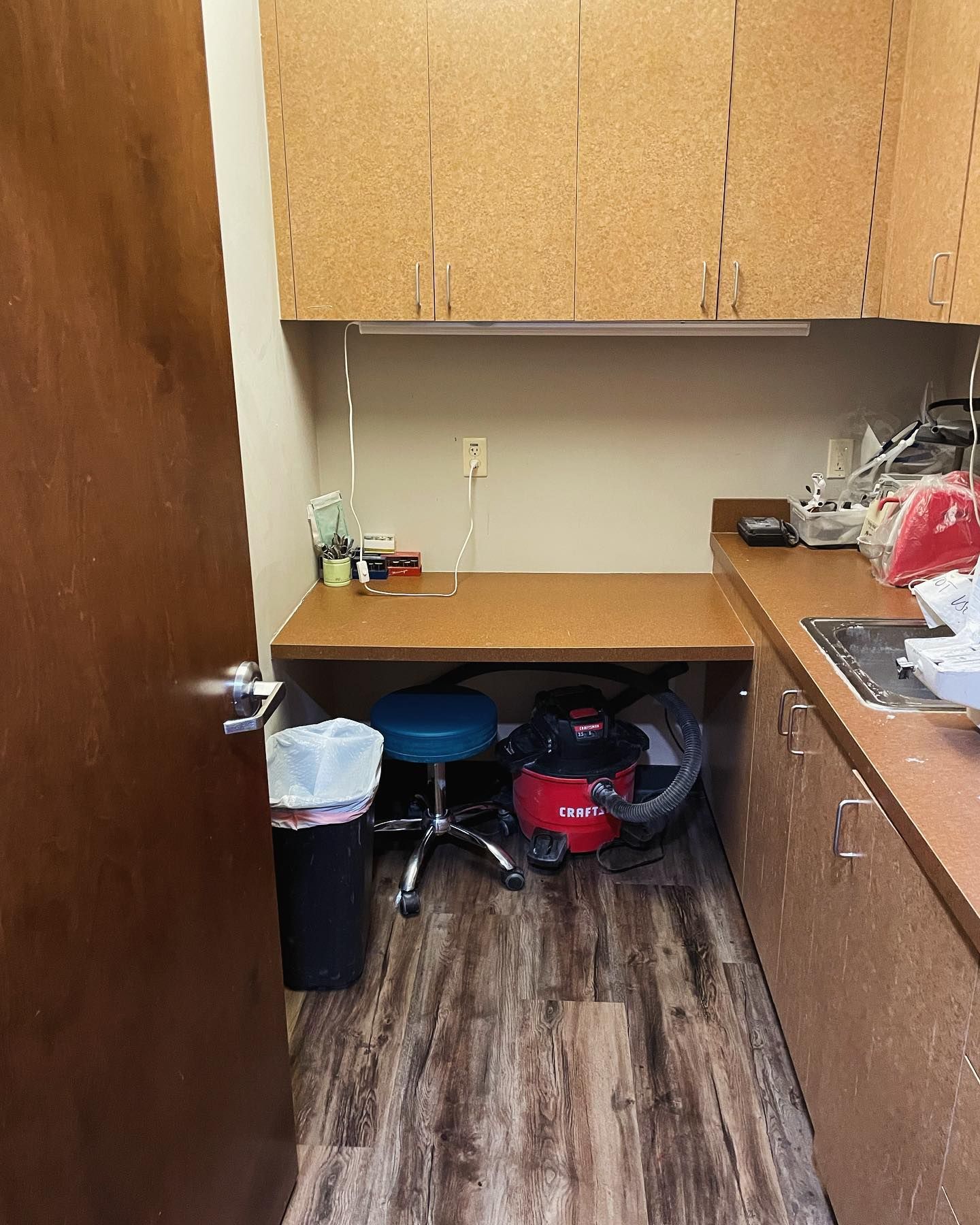 Small room with a desk, cabinets, vacuum cleaner, trash can, and blue stool.