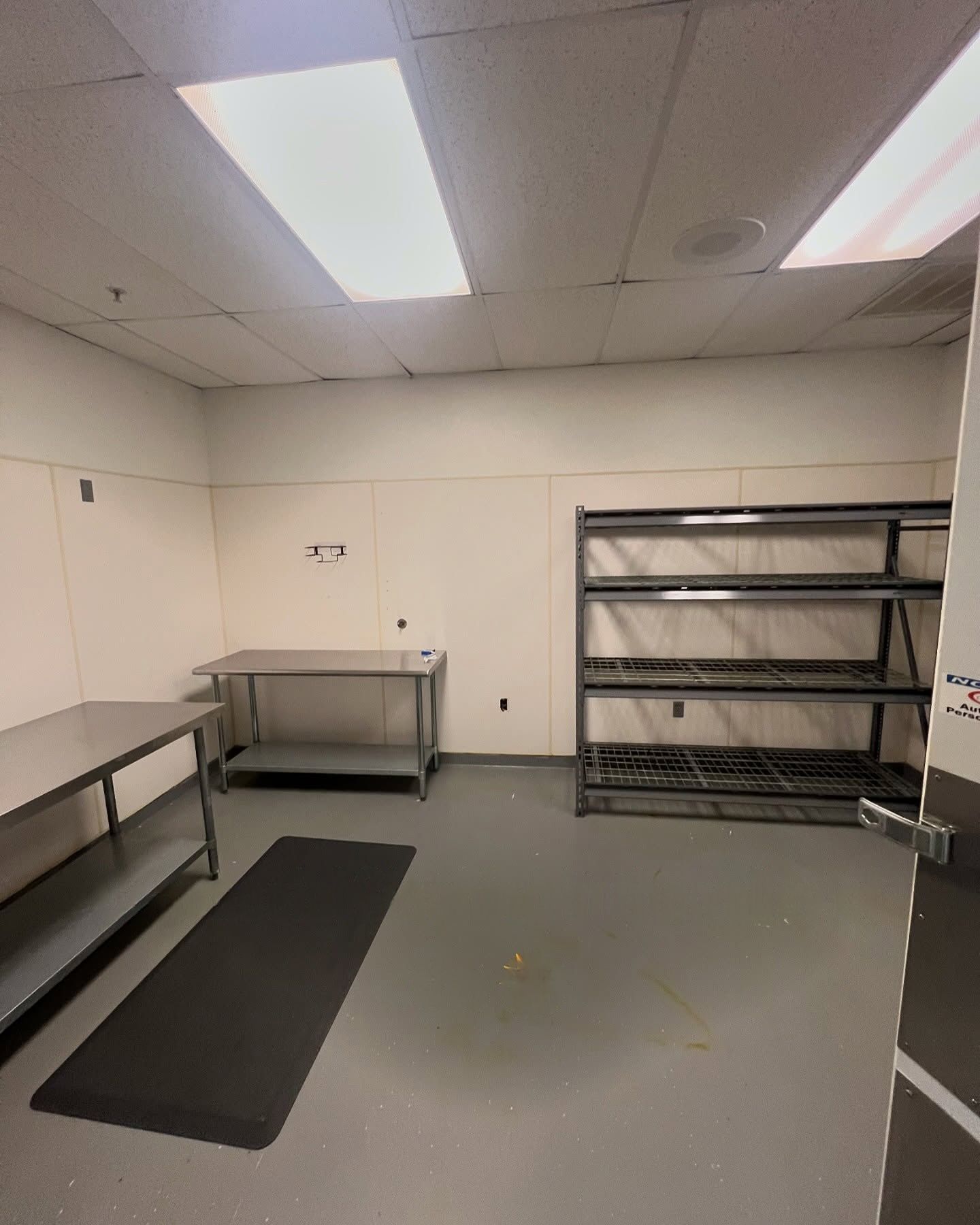 Empty commercial kitchen with stainless steel tables, shelving, gray floor, and overhead fluorescent lights.