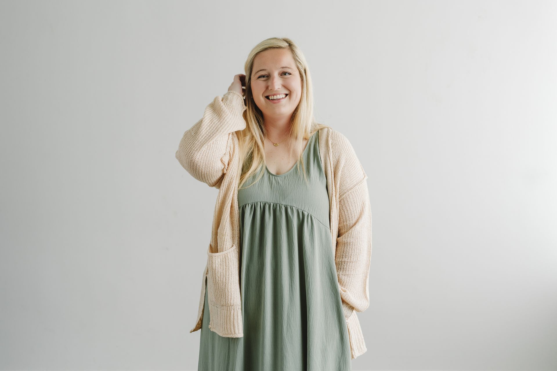 A woman in a green dress and cardigan is standing in front of a white wall.