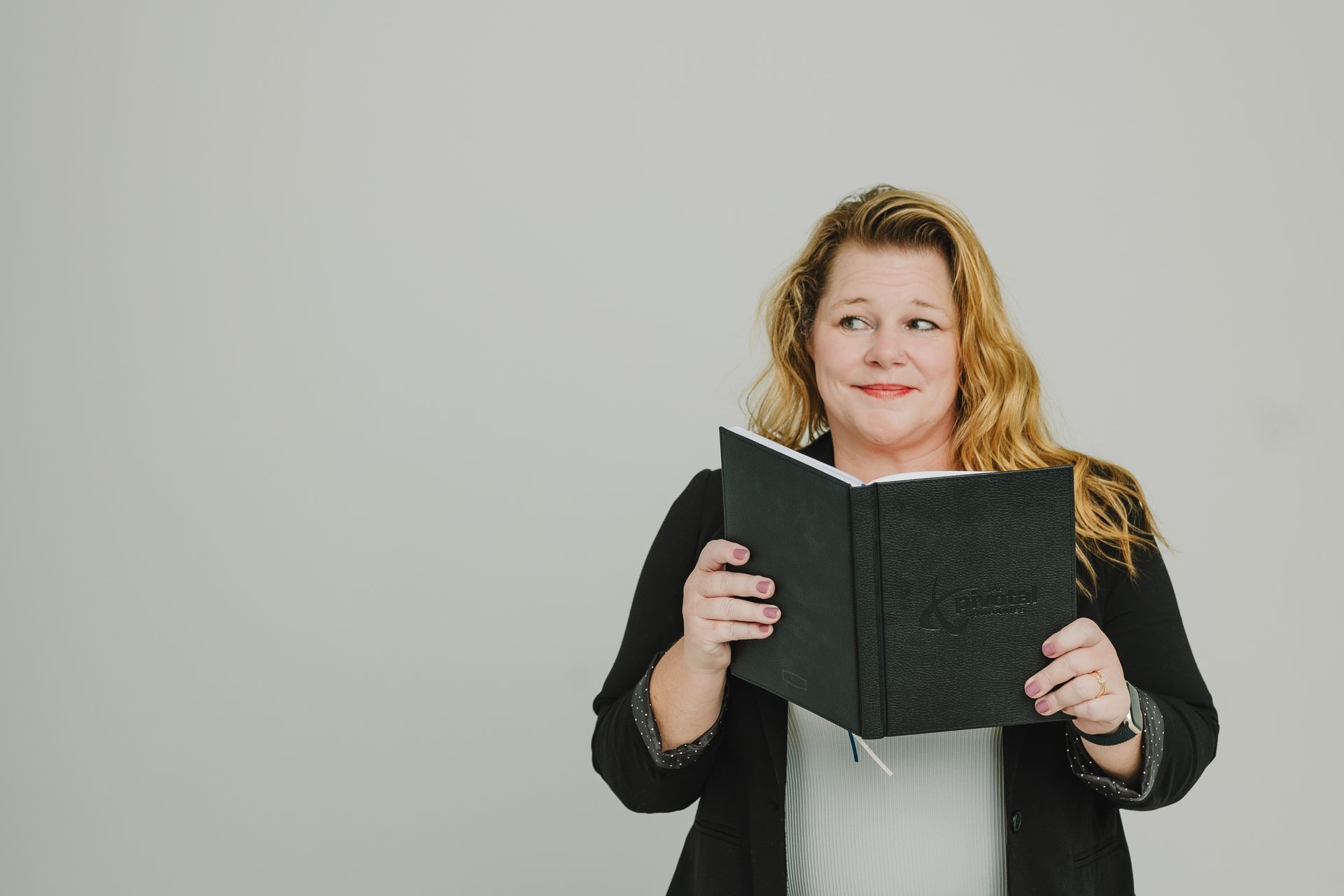 Woman with blonde hair holding a book, looking to the side, smiling. Against a plain background.
