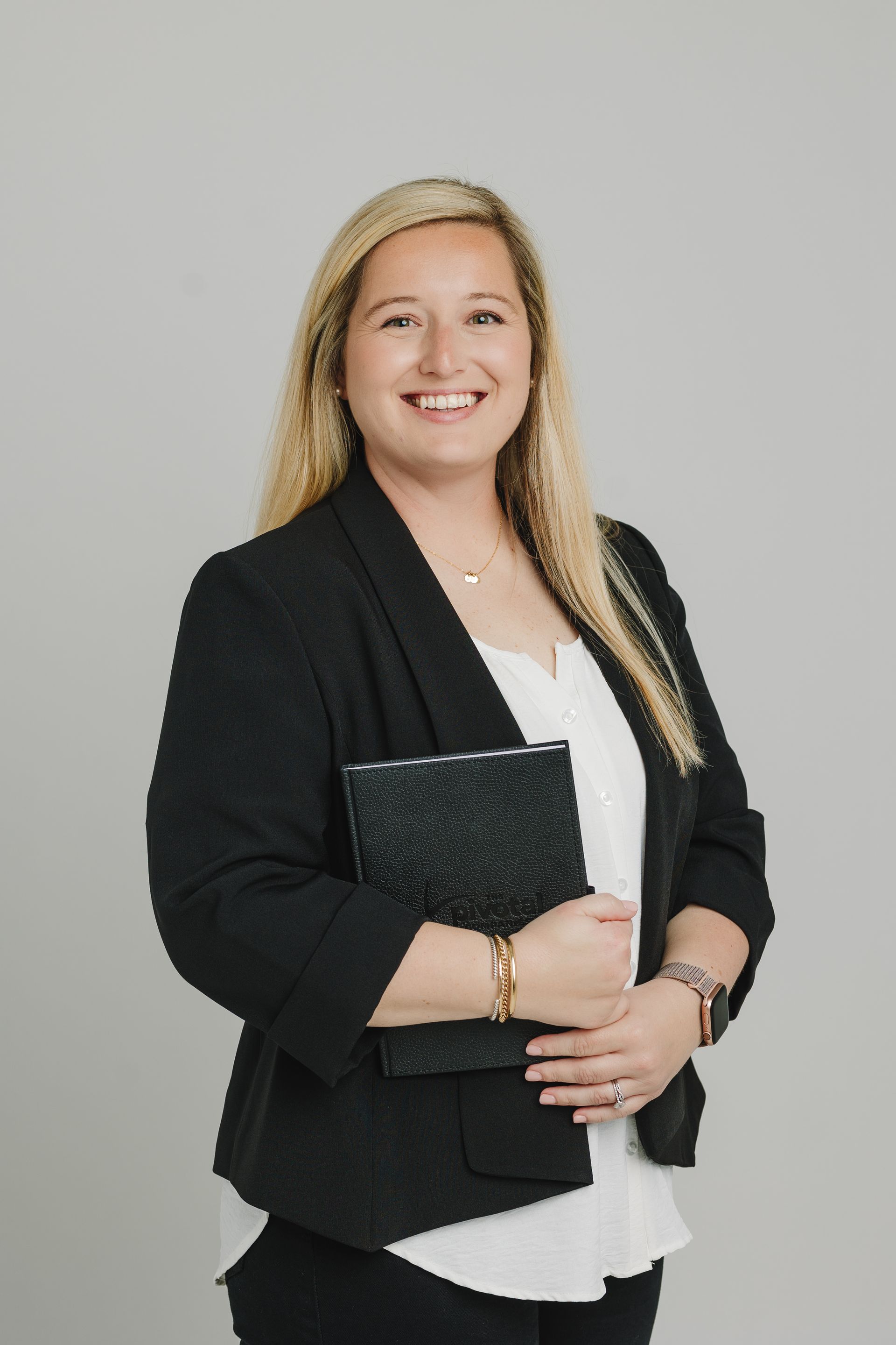 Blonde woman in black blazer, white shirt, holding a folder, smiling against a gray background.