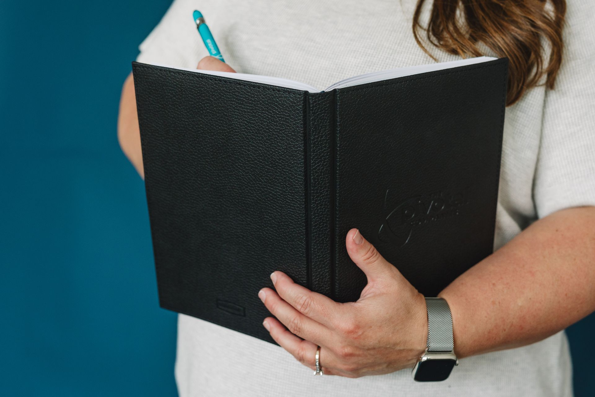Woman holding a black notebook, writing with a blue pen, blue background.