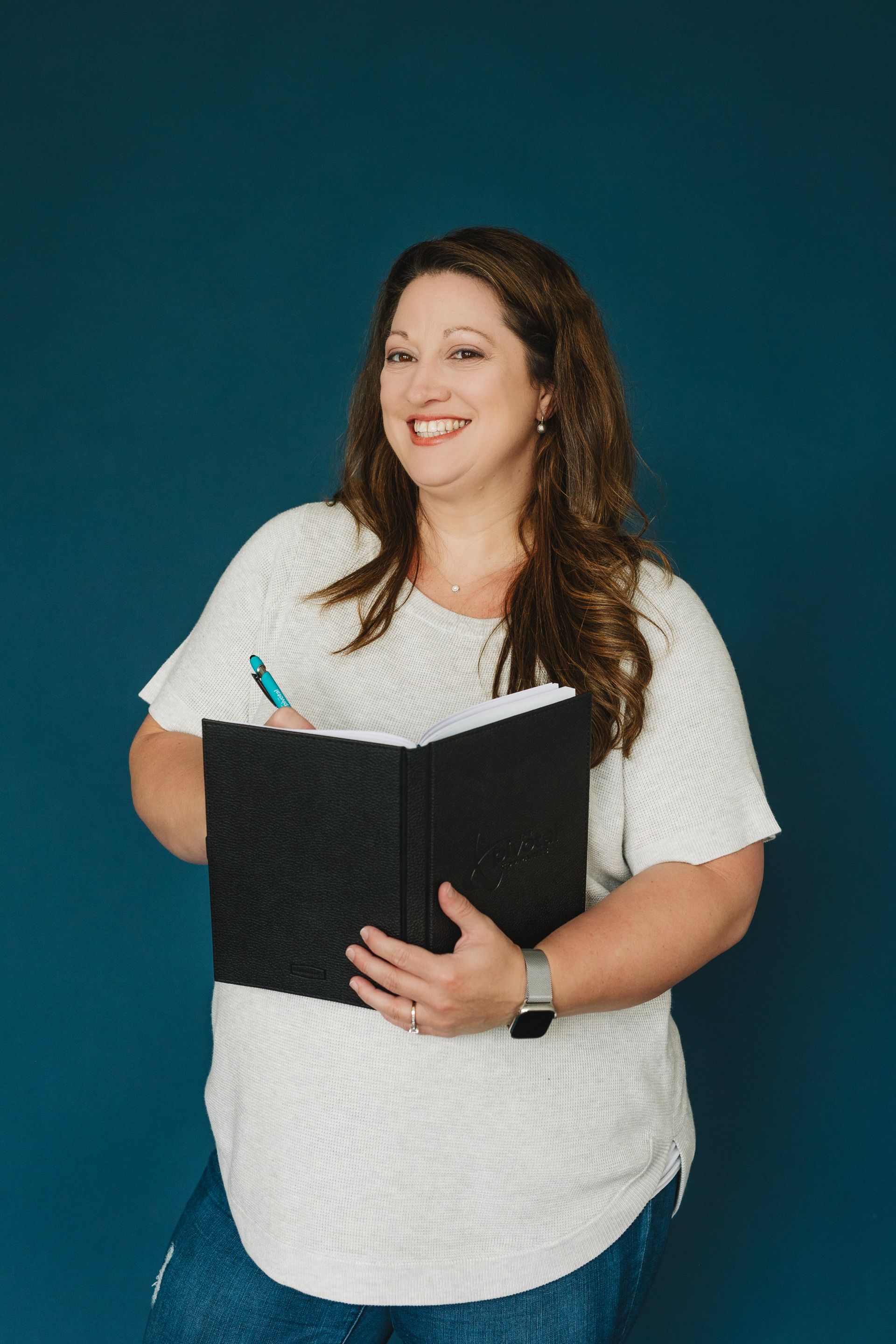 A woman in a white shirt is holding a black book and a pen.