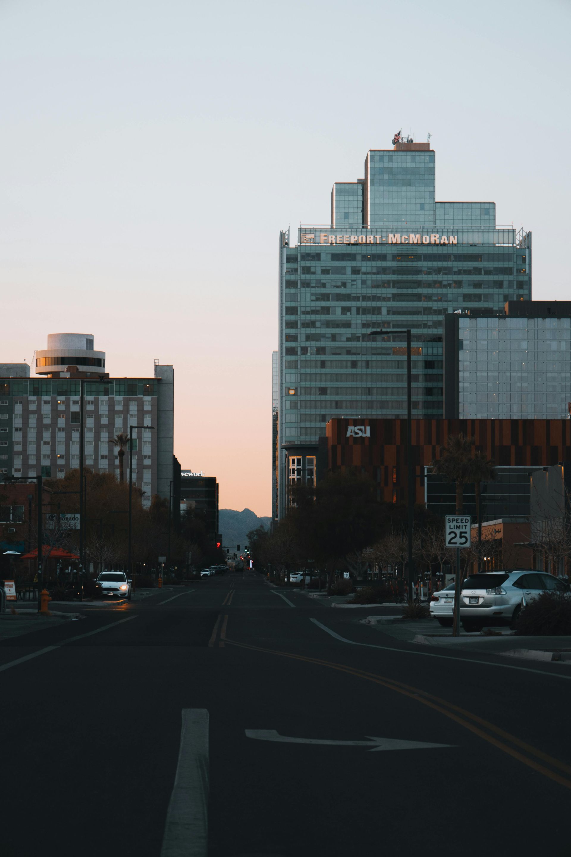 City street with tall buildings, cars, and streetlights against a dusky sky.
