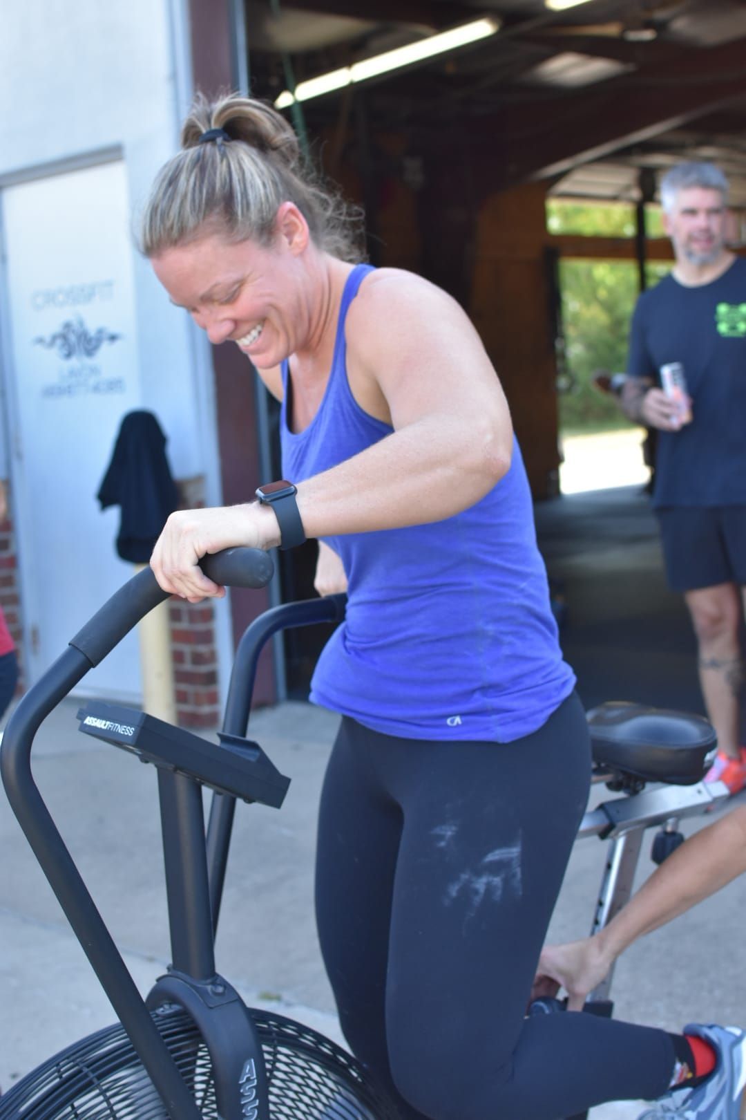 Woman in blue tank top and black leggings on a stationary bike at a Lavon, TX gym, smiling. Outdoors.