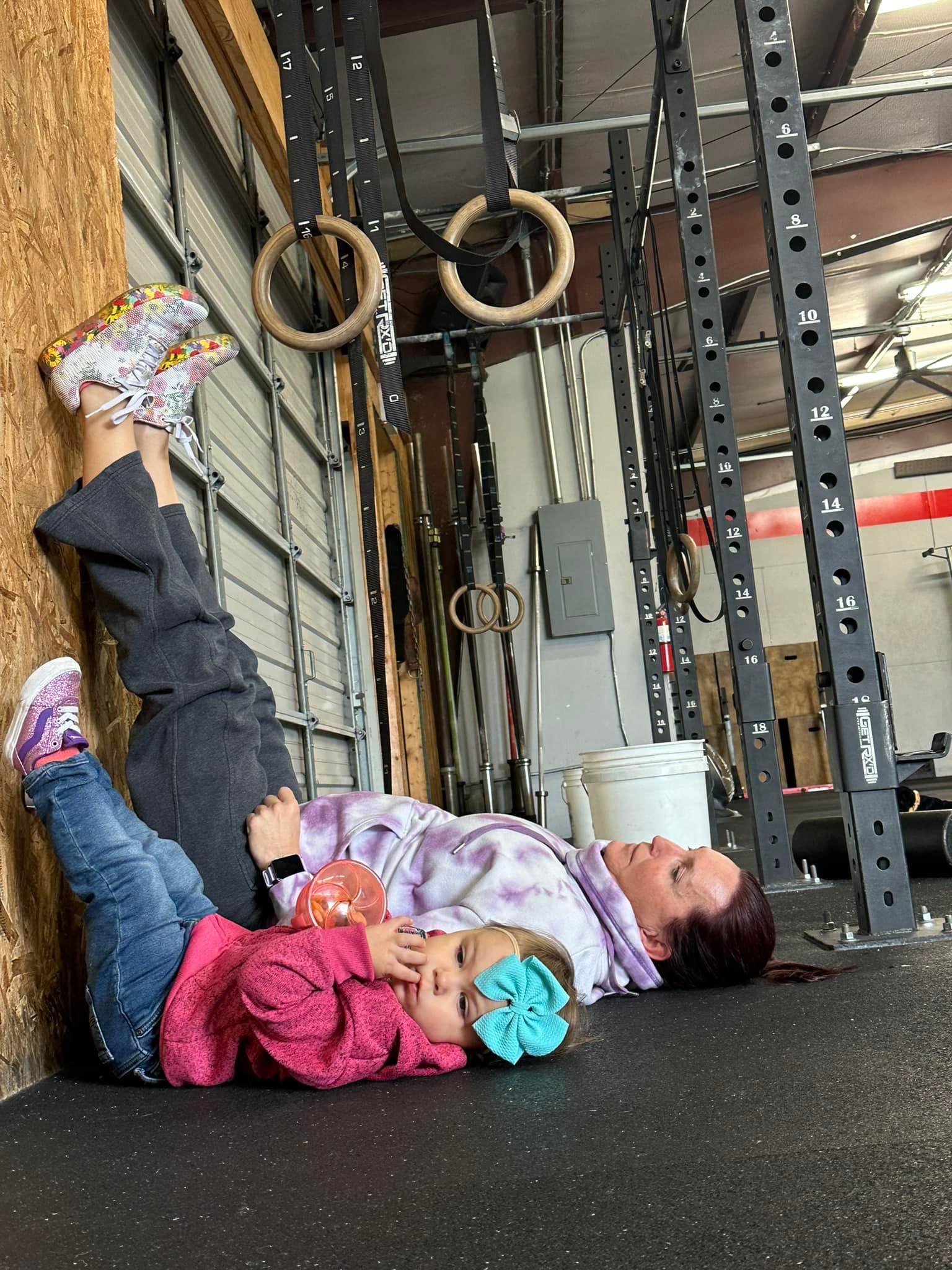 Woman and child resting on the floor at a gym in Lavon, TX, feet up on wall. Gym rings and equipment visible.
