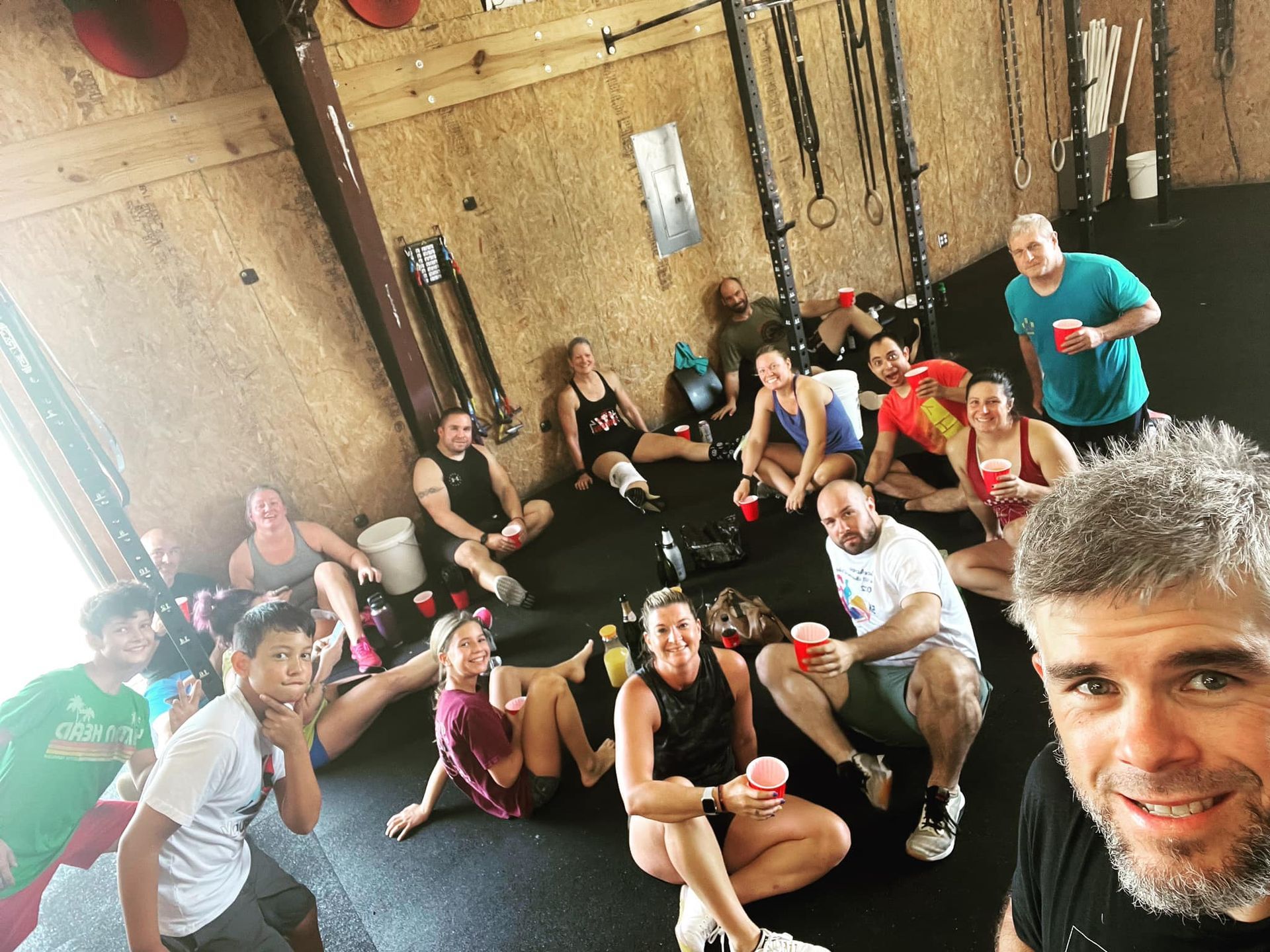 Group of people relaxing on a gym floor after a workout, some holding red cups.