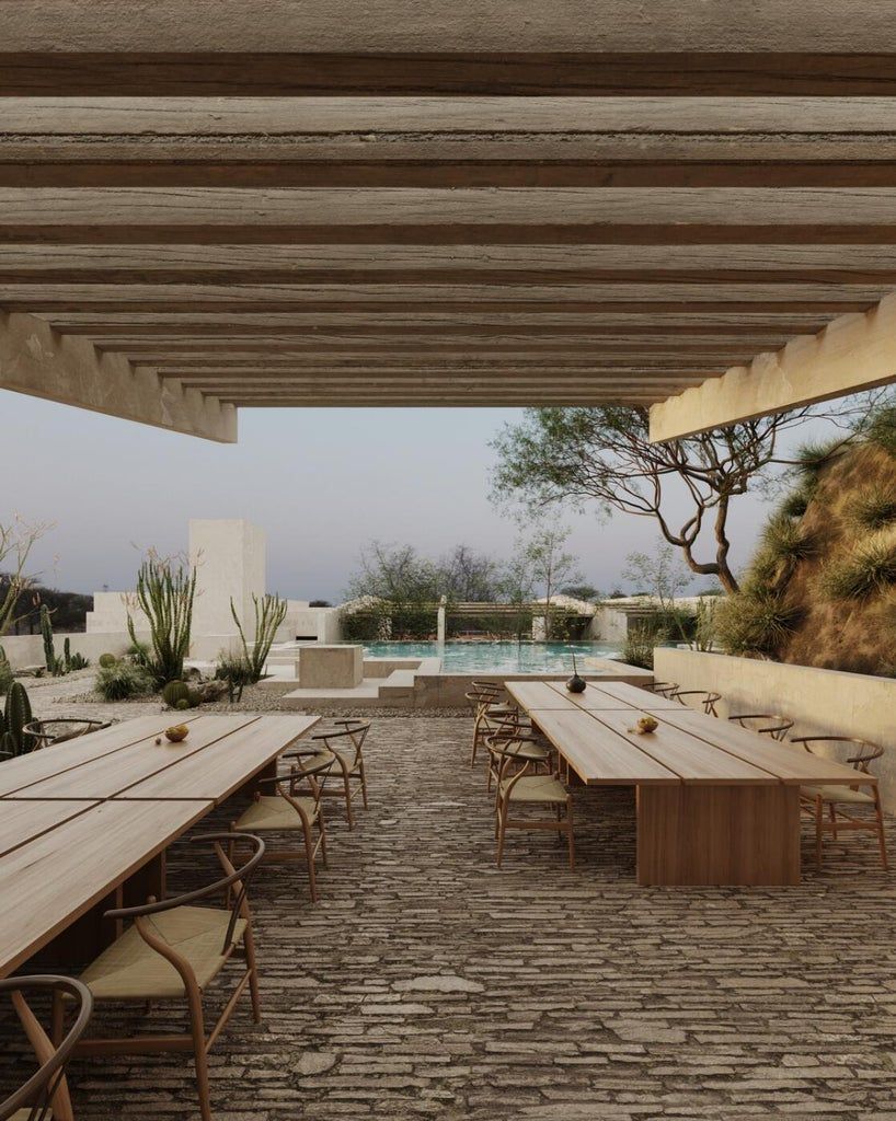 Outdoor dining area with long wooden tables, chairs, and a pool in the background, under a wooden pergola.