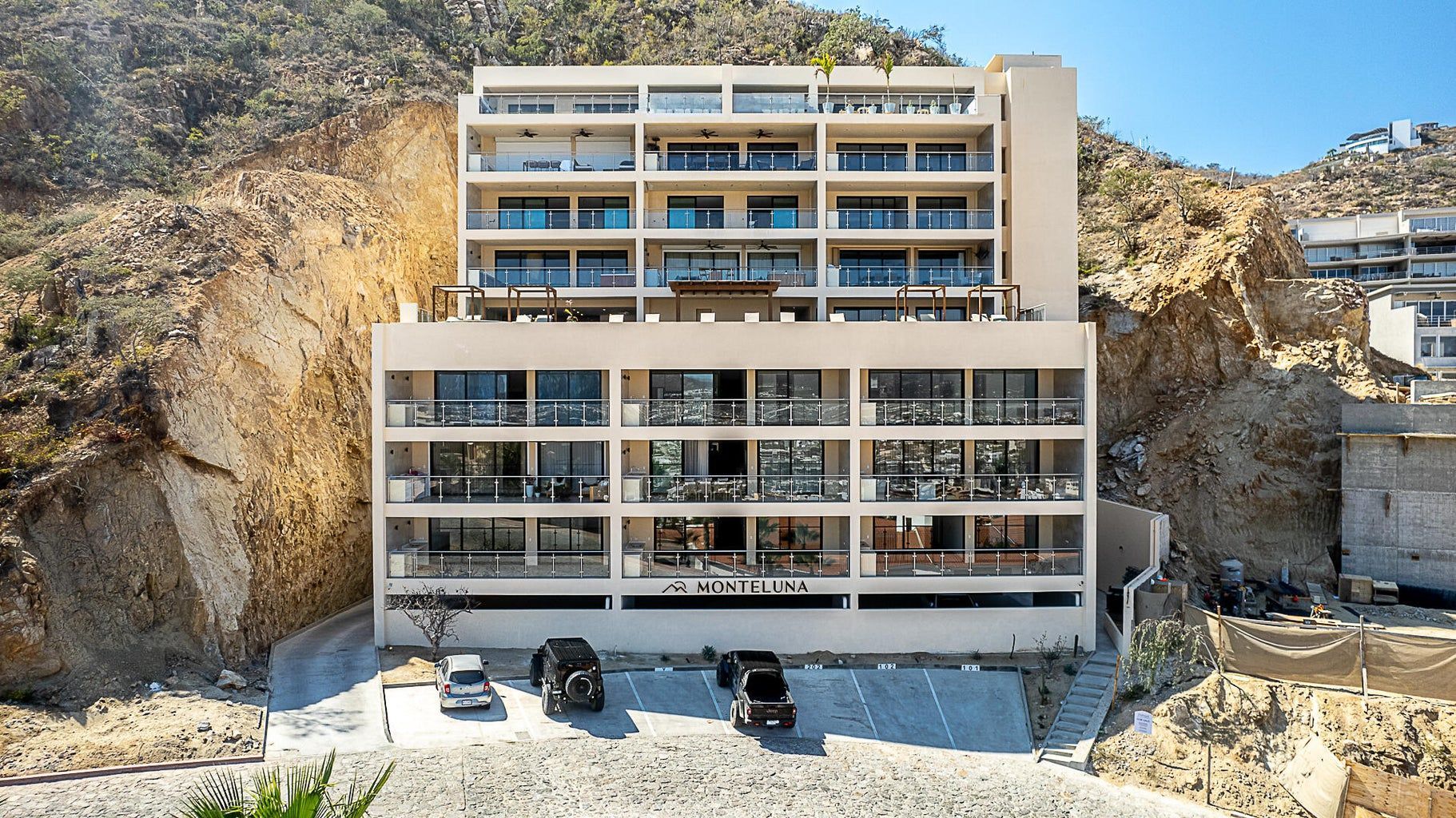 Modern multi-story building cut into a hillside, with balconies and cars parked out front.
