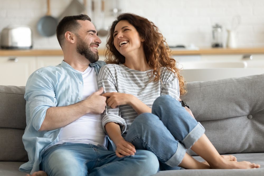 Couple laughing on a sofa in a home setting. Man in blue shirt, woman in striped shirt.