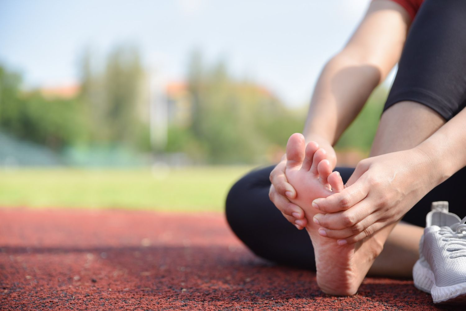 Person sitting on a track, holding foot. Red track, green background, gray shoes.