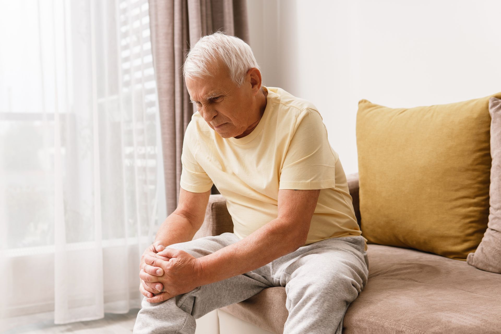 An elderly man is sitting on a couch holding his knee in pain.